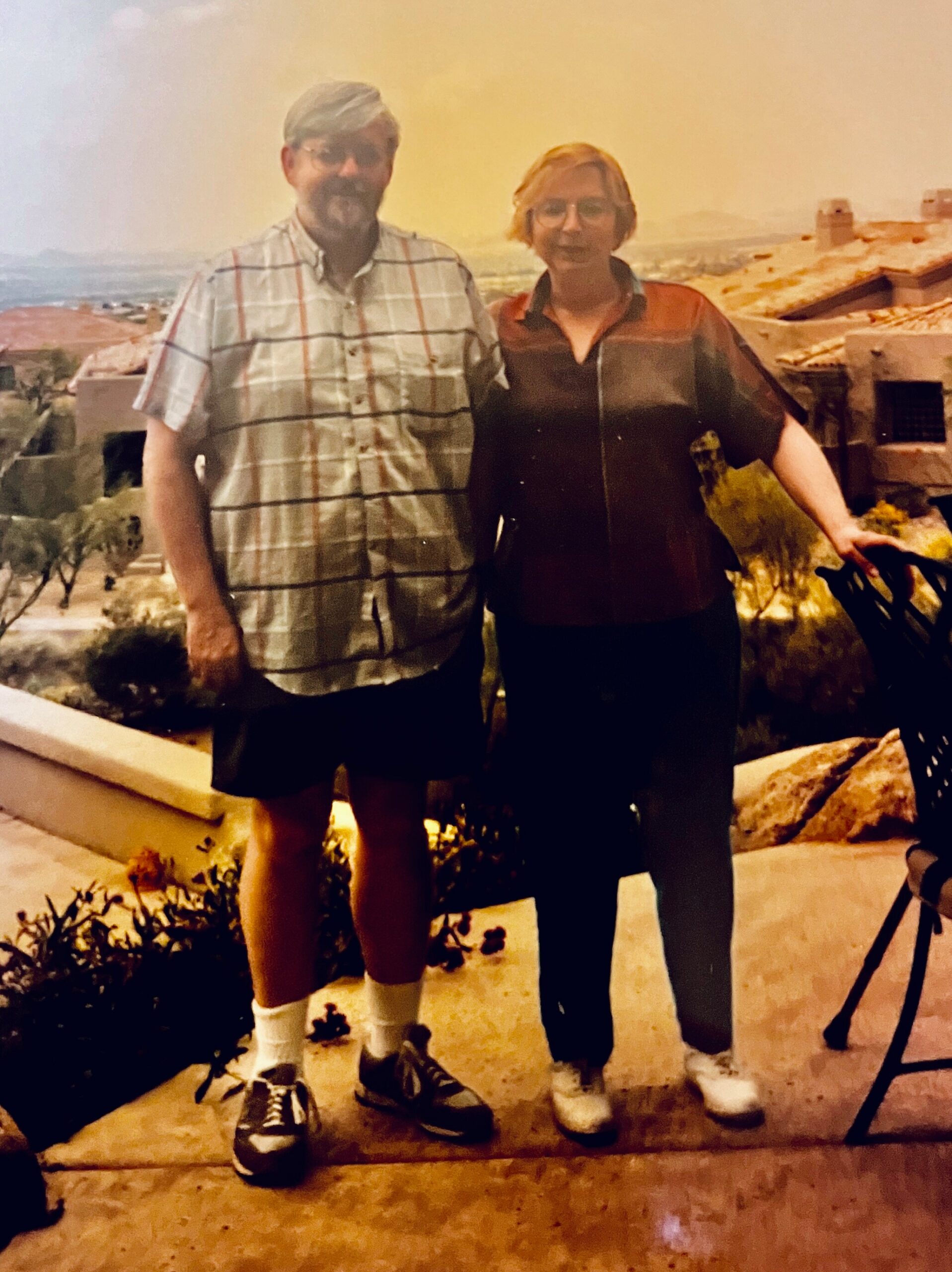 Older couple posing together on a sunlit terrace with a desert town in the background at sunset.