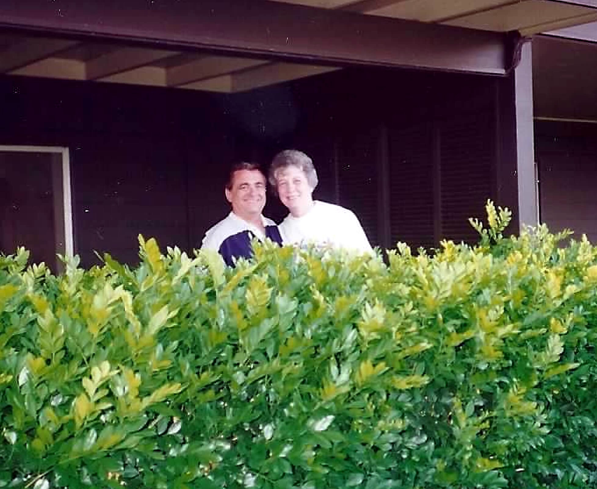 Two people smiling and posing behind a green hedge outdoors.