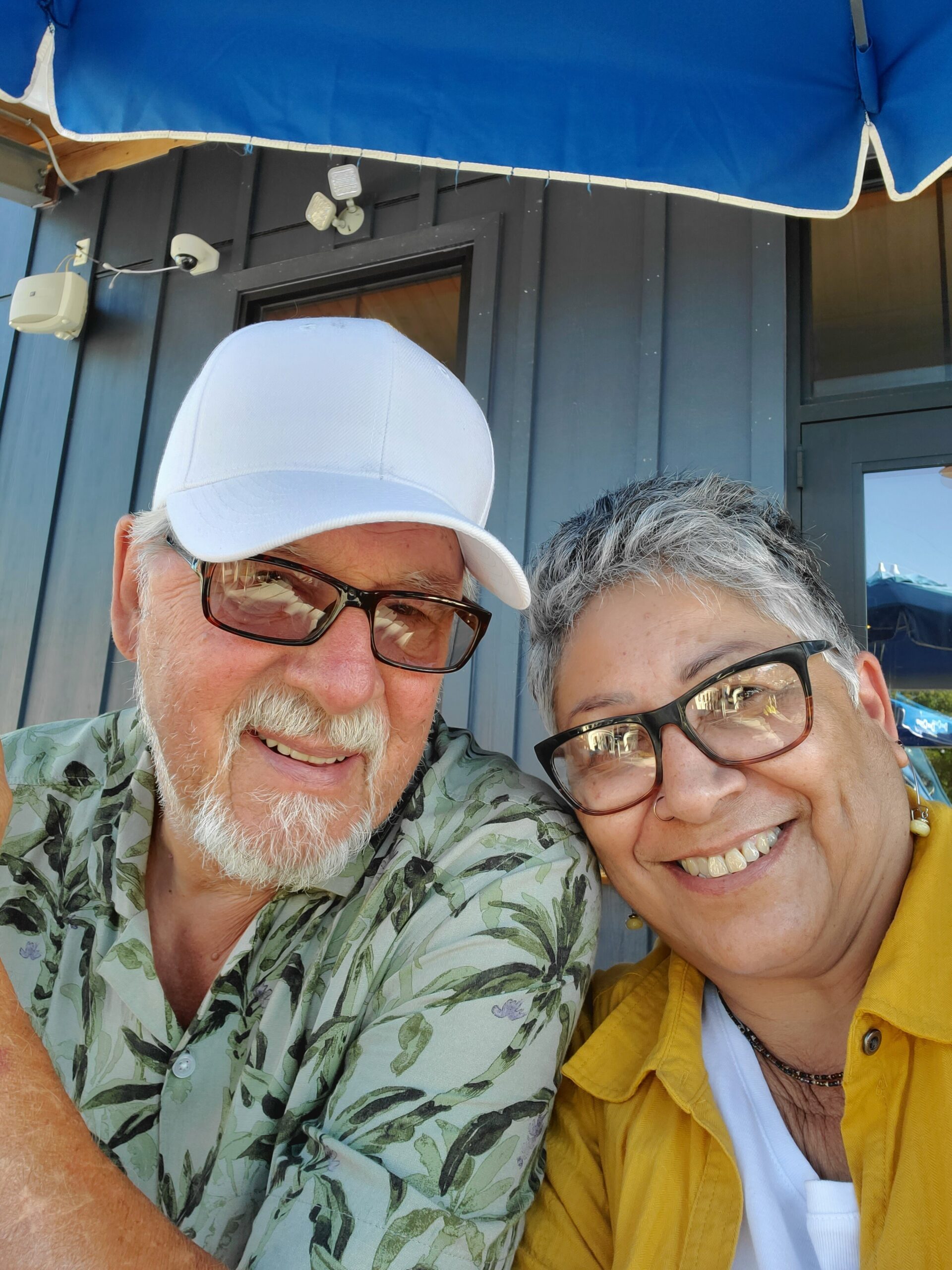 Two smiling seniors pose for a selfie outside a blue building; man wears a white cap and patterned shirt, woman wears glasses and a yellow jacket.
