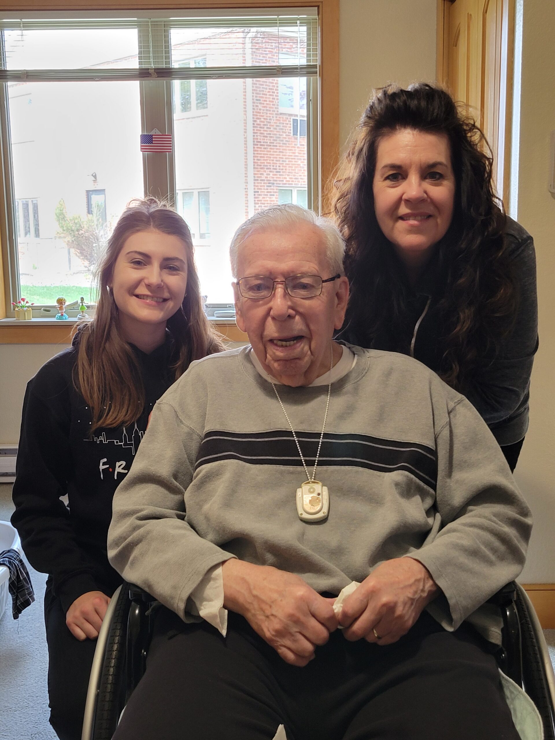 Elderly man in a wheelchair sits center, flanked by two smiling women indoors near a window.