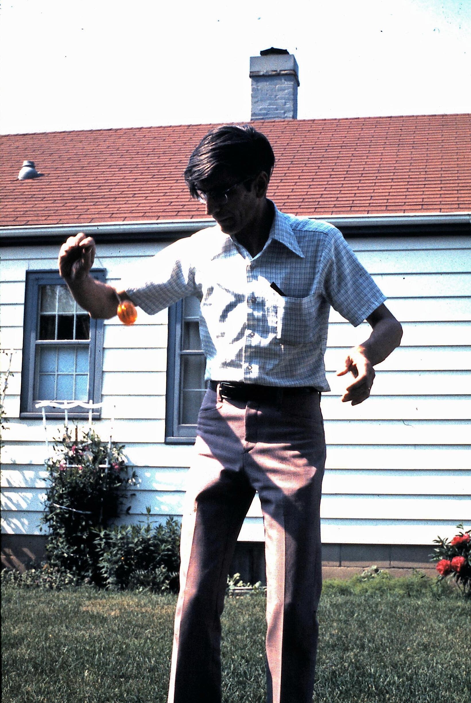 Man wearing a short-sleeve checkered shirt and trousers, outdoors in a backyard, juggling an orange ball in one hand.