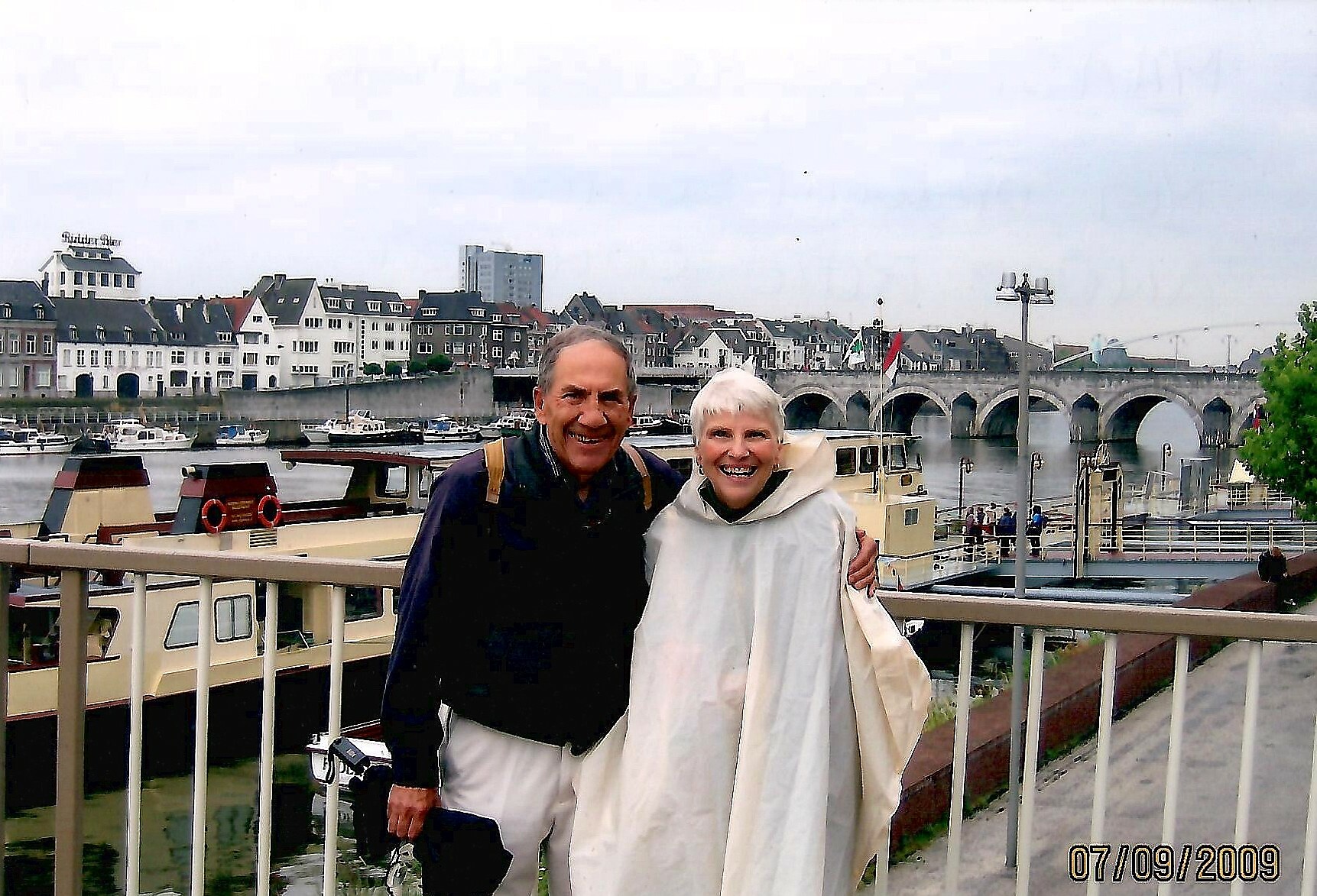 Older couple smiling, standing on a railing overlooking a harbor with boats and a stone-arched bridge in a European town. (07/09/2009)