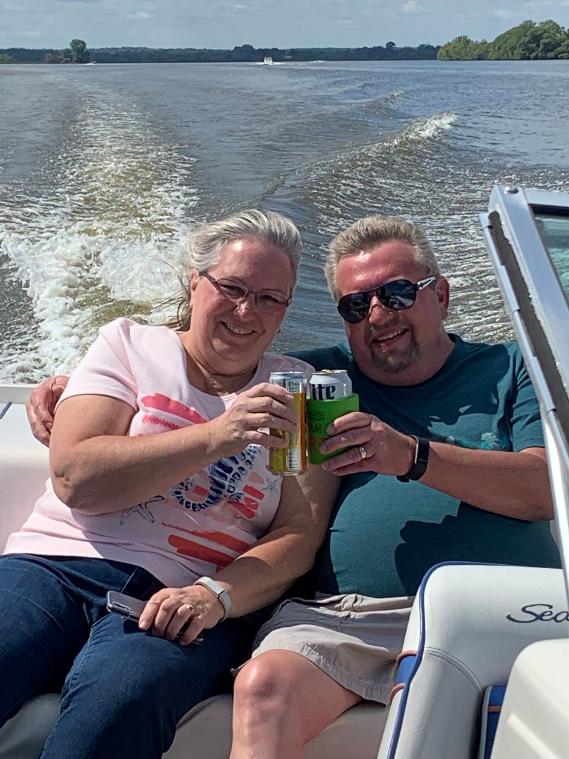 Two adults on a boat toasting with beer cans, smiles at the camera, water and wake behind them on a sunny day