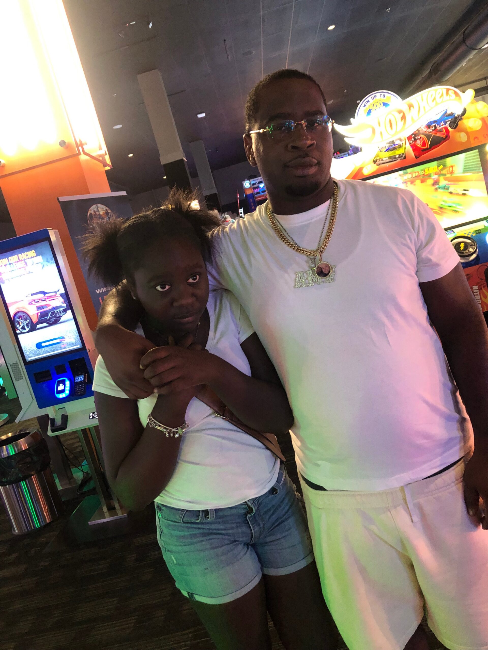 Man and girl posing closely in a bright arcade, the man in a white shirt and gold chain next to an arcade racing game setup with a Hot Wheels sign in the background.