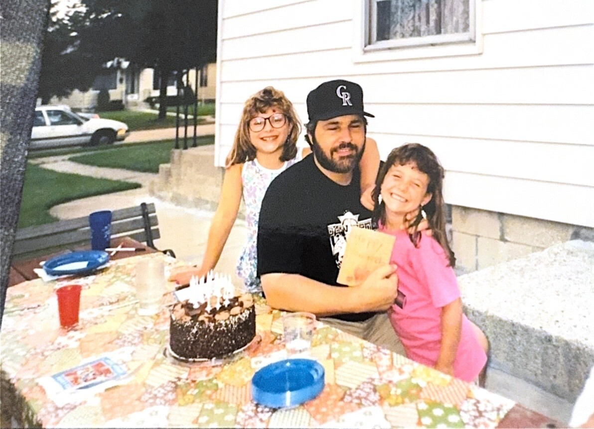 Man with beard and two girls posing at a birthday party outdoors; cake with candles on a patterned tablecloth in front of them.