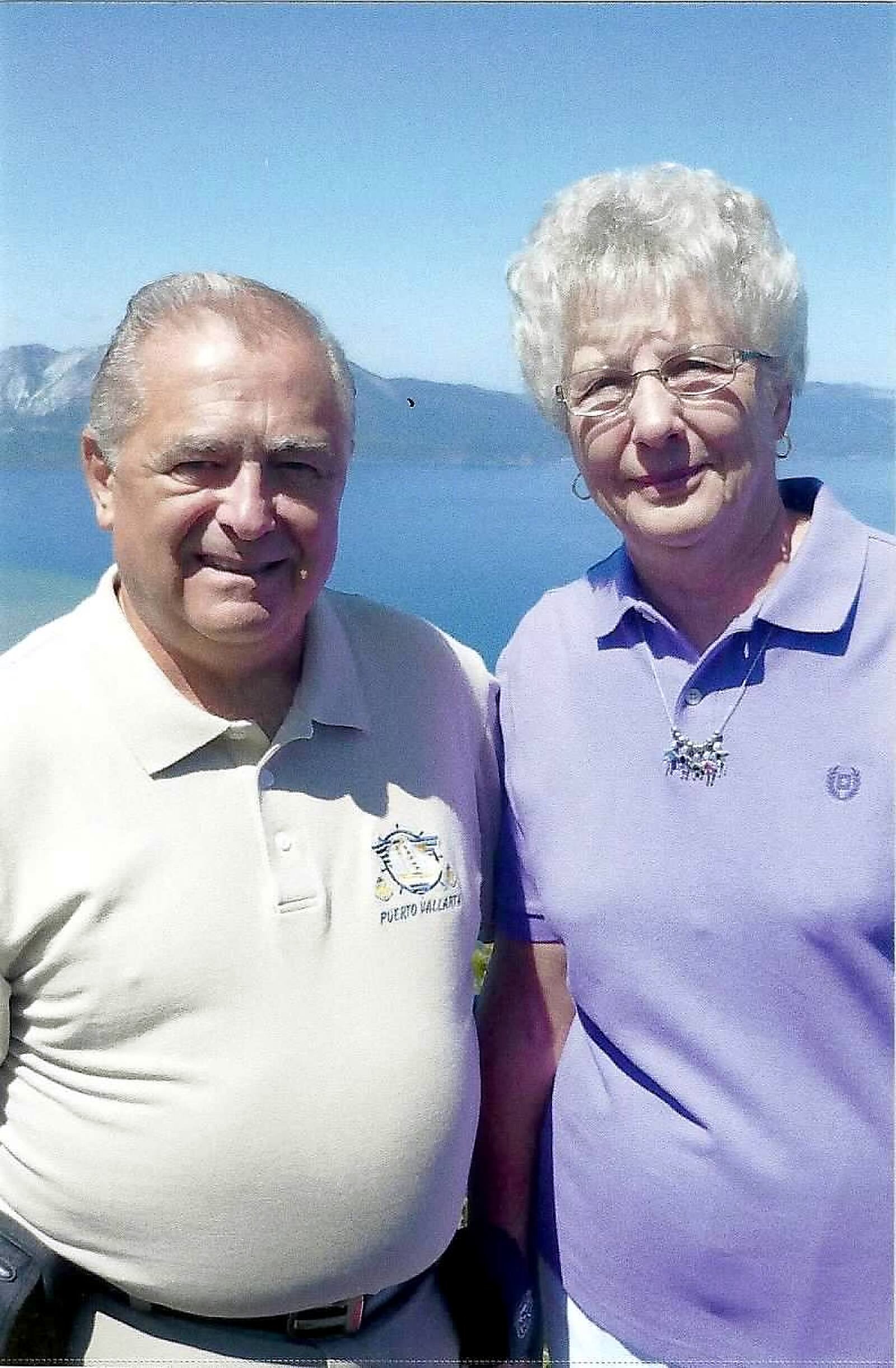 Two smiling older adults posing outdoors with a blue lake and mountains in the background.