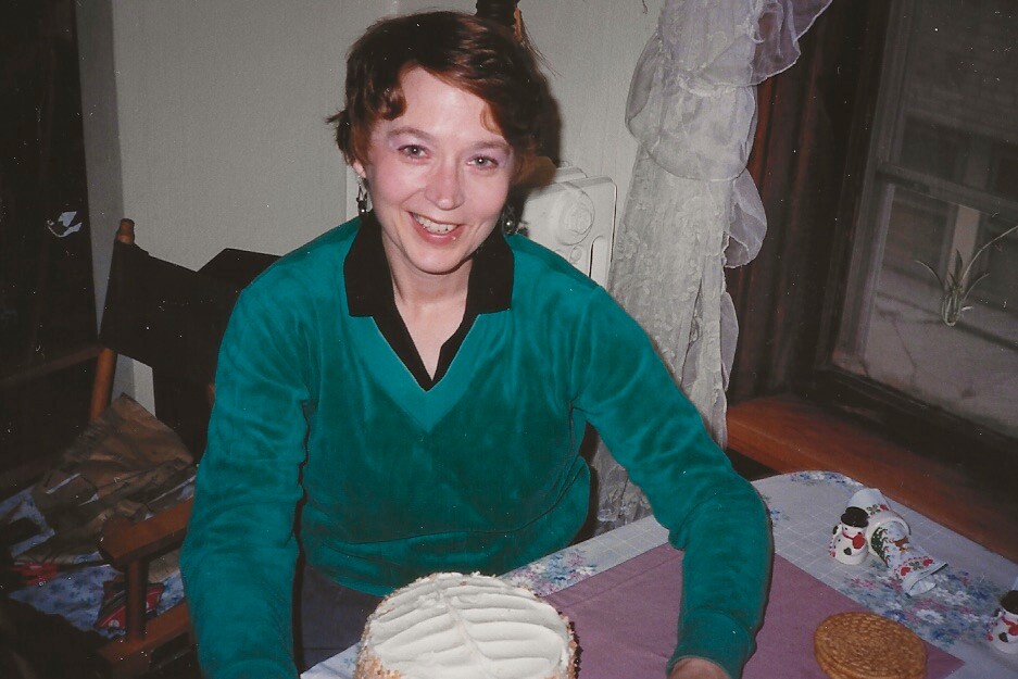 Woman in a teal top sits at a table with a frosted cake, smiling at the camera in a cozy room.