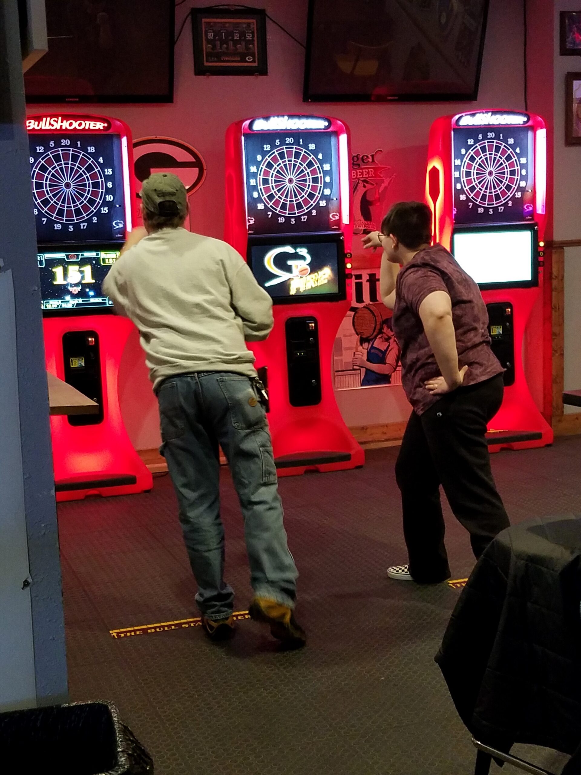 Two people stand at red electronic dart machines in a dim arcade, focusing on their darts at the boards.