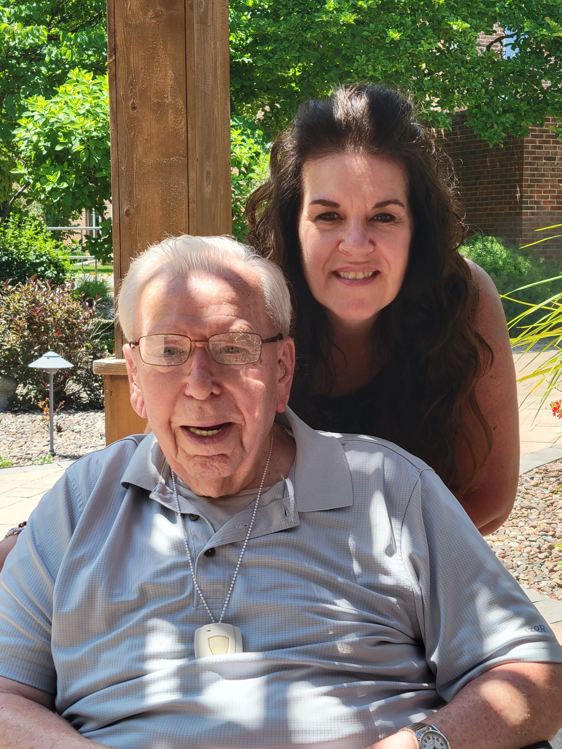 An elderly man in a light striped polo sits in front, with a smiling woman behind him, outdoors in a sunny garden.