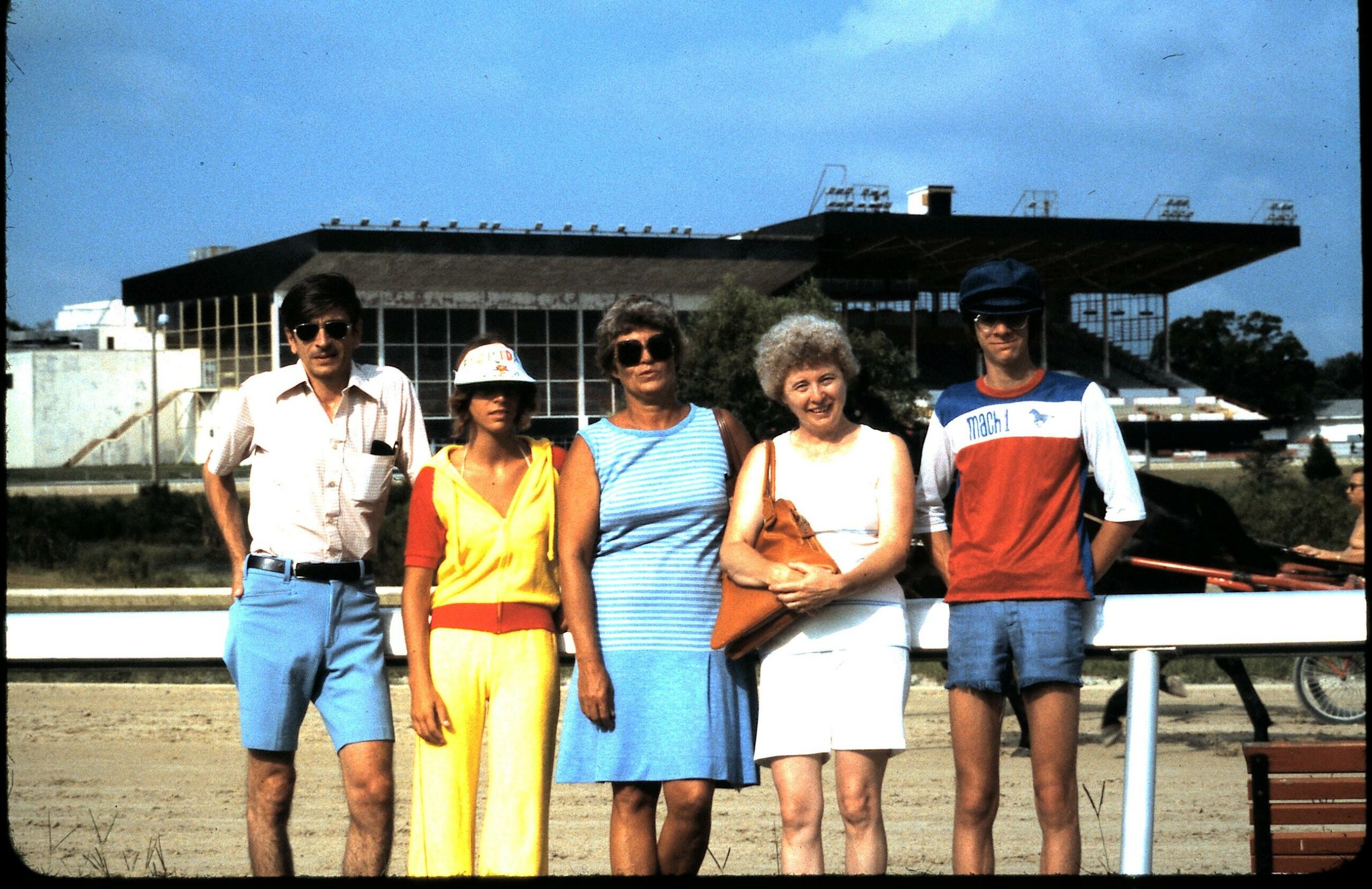 Five adults pose together outdoors in front of a large stadium bleacher structure on a sunny day.