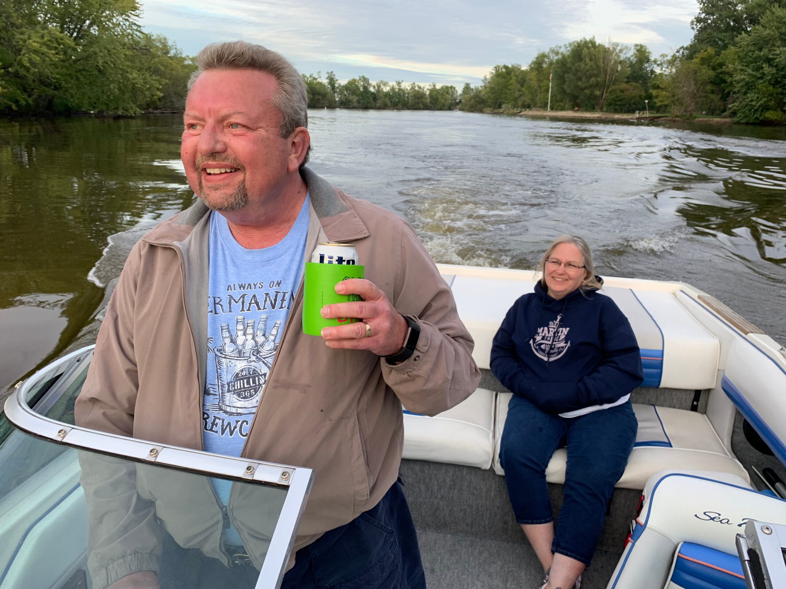 Man in a beige jacket smiling while holding a can on a boat, with a woman sitting in the background by the rail. The river and trees are in view.