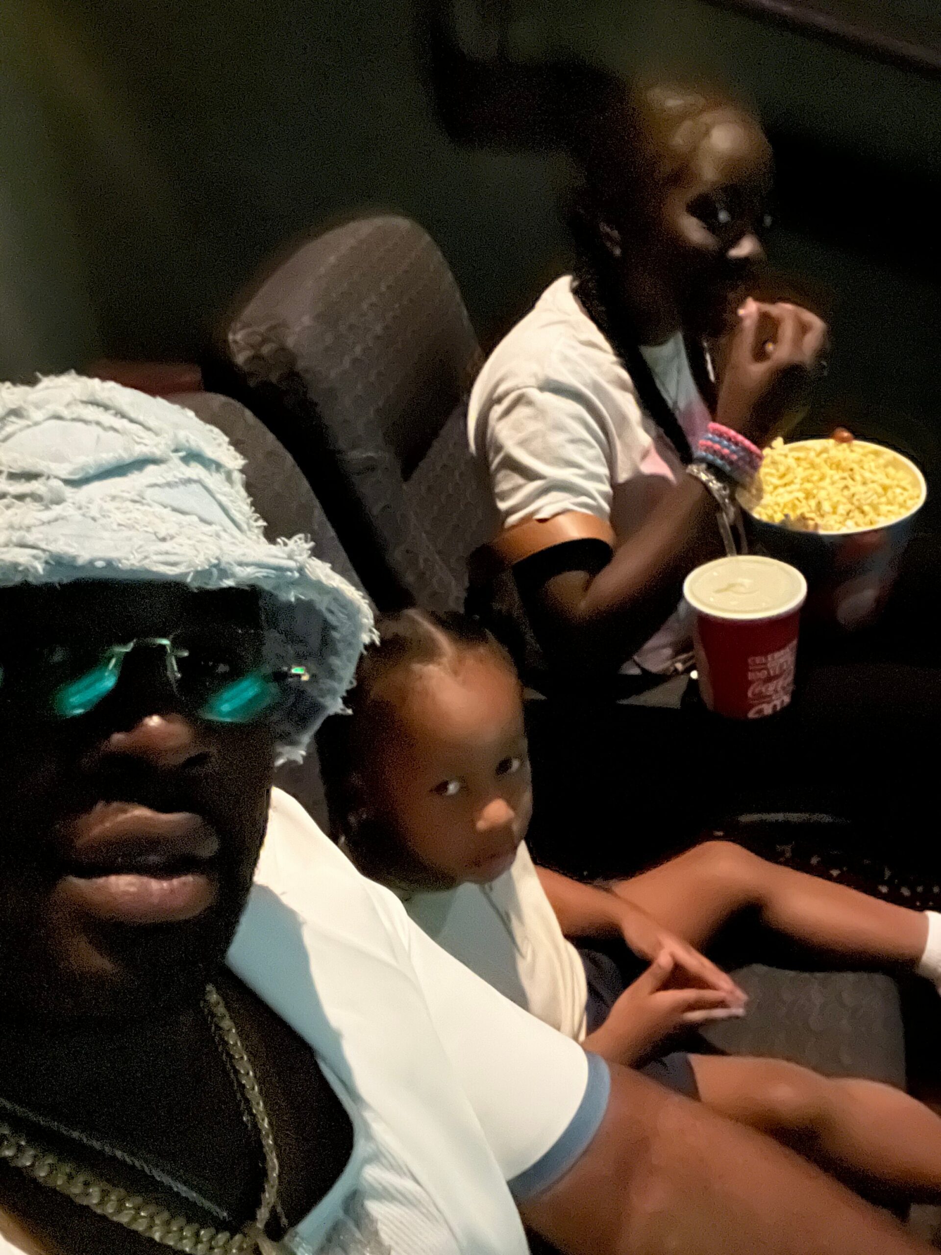 People seated in a dark movie theater; foreground person wears a light blue outfit and a textured headwrap, a child looks toward the camera, others snack on popcorn.