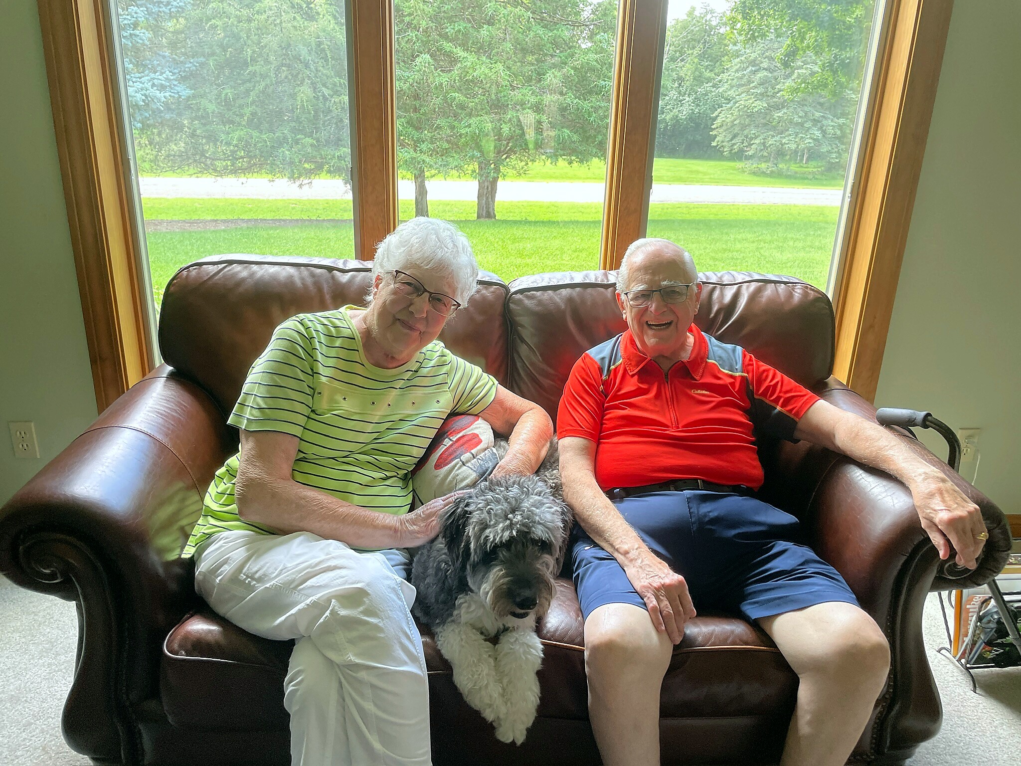 Two seniors sit on a brown leather couch with a fluffy gray dog between them, smiling at the camera in a bright living room. Outside, green trees and lawn are visible through large windows.