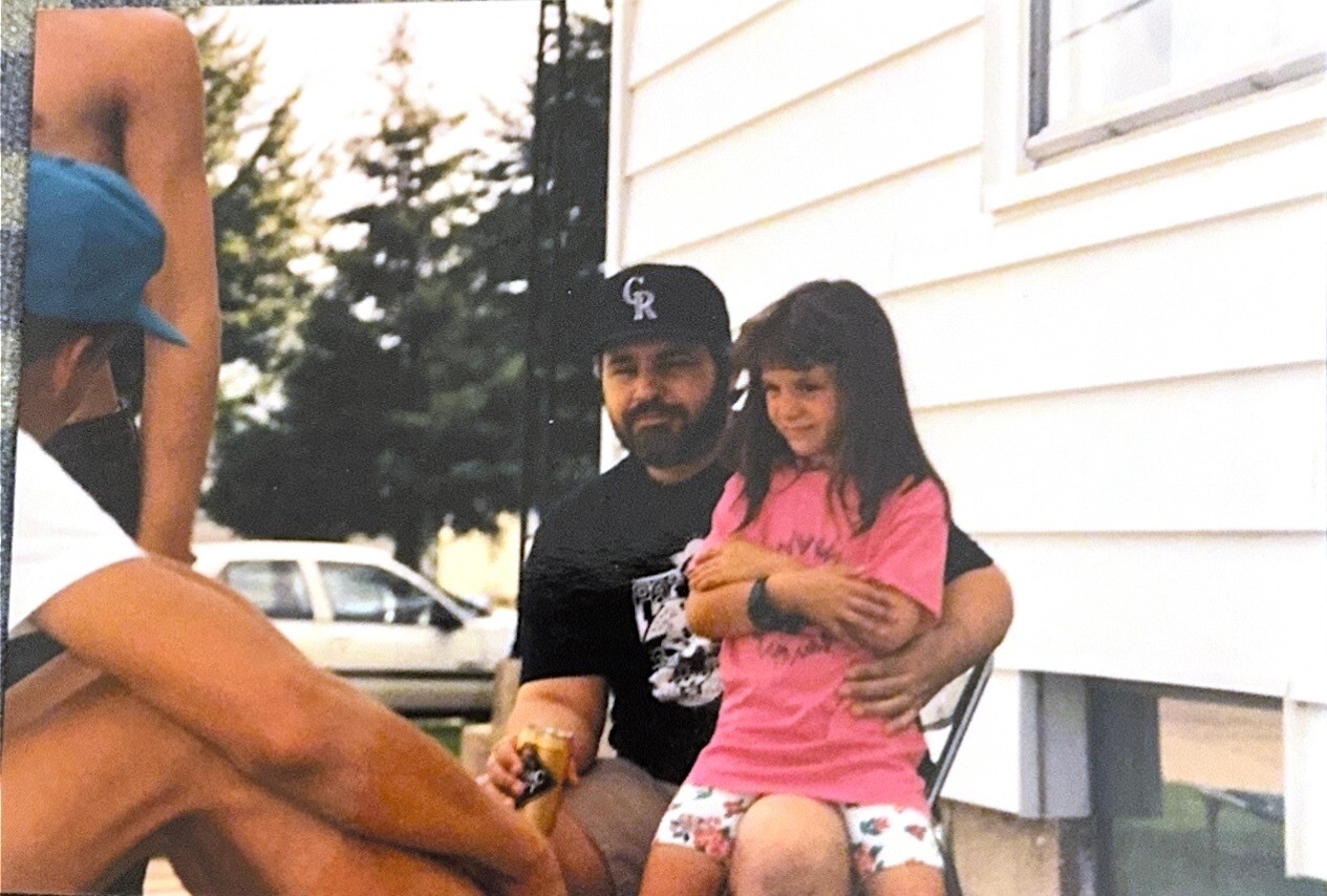 Bearded man in a black t‑shirt and cap sits on a porch, holding a young girl in a pink shirt and floral shorts.