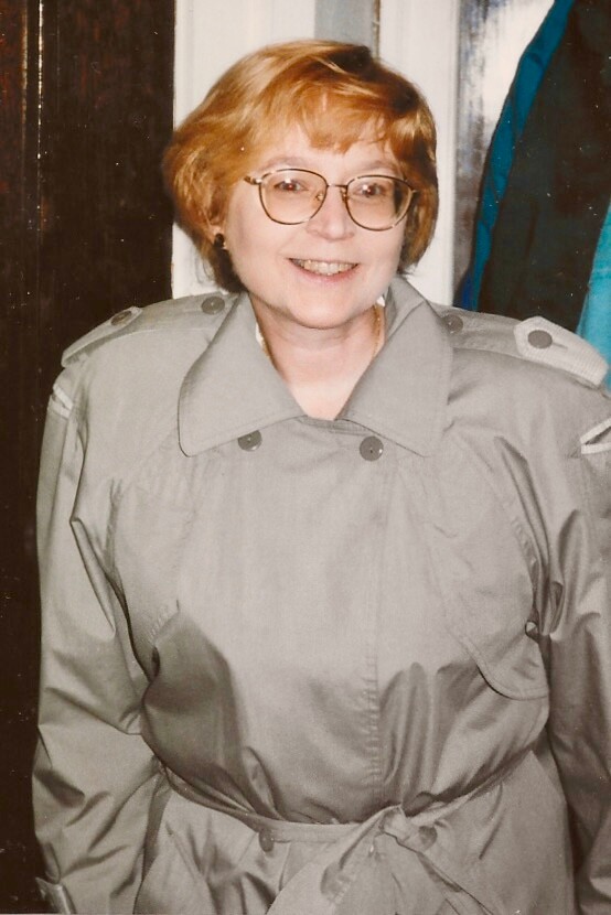 Smiling woman with short red hair and glasses wearing a light-gray trench coat indoors.