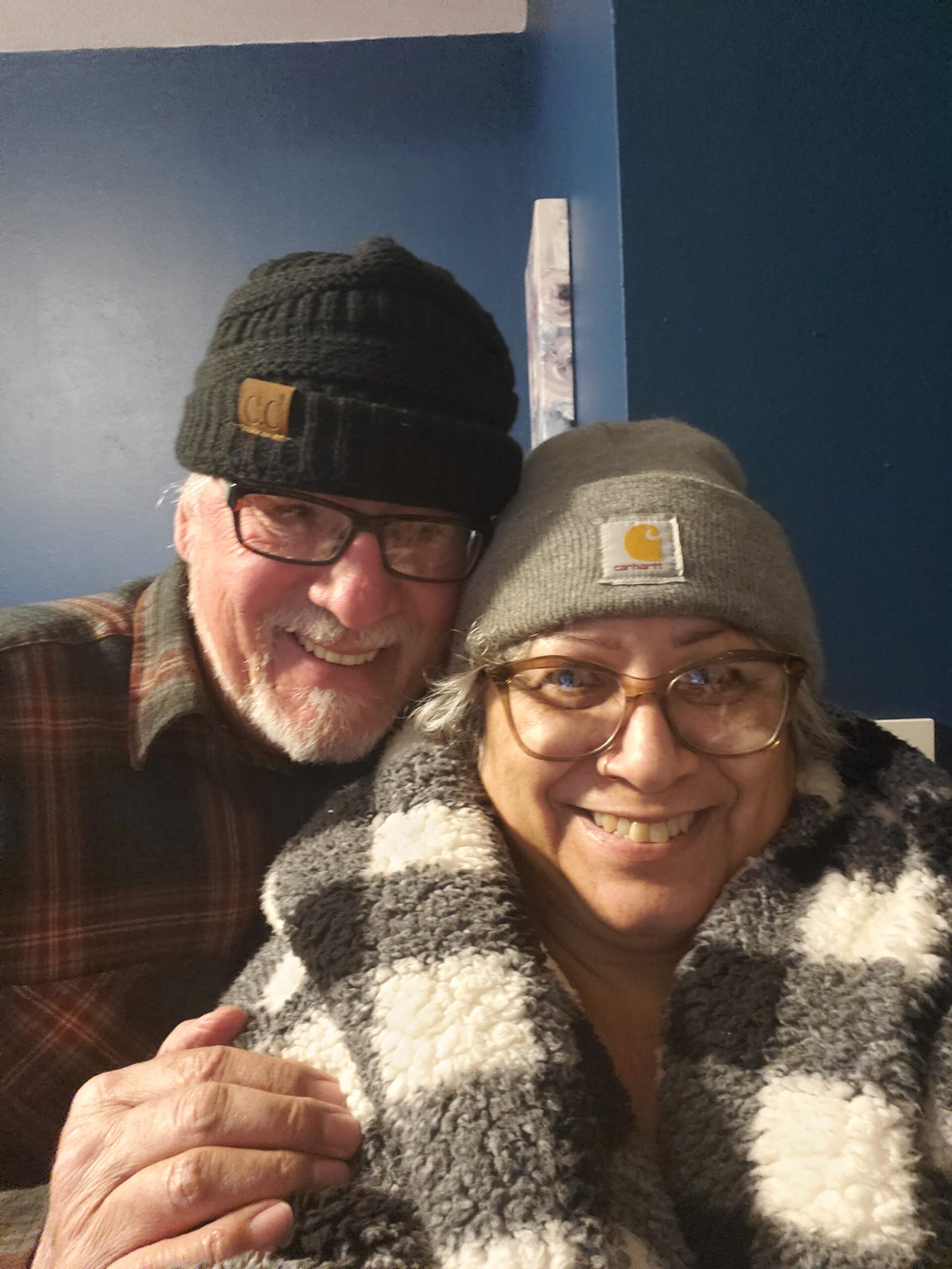 Smiling elderly couple wearing warm hats indoors: man in a black knit beanie and glasses, woman in a gray Carhartt beanie and fuzzy coat.