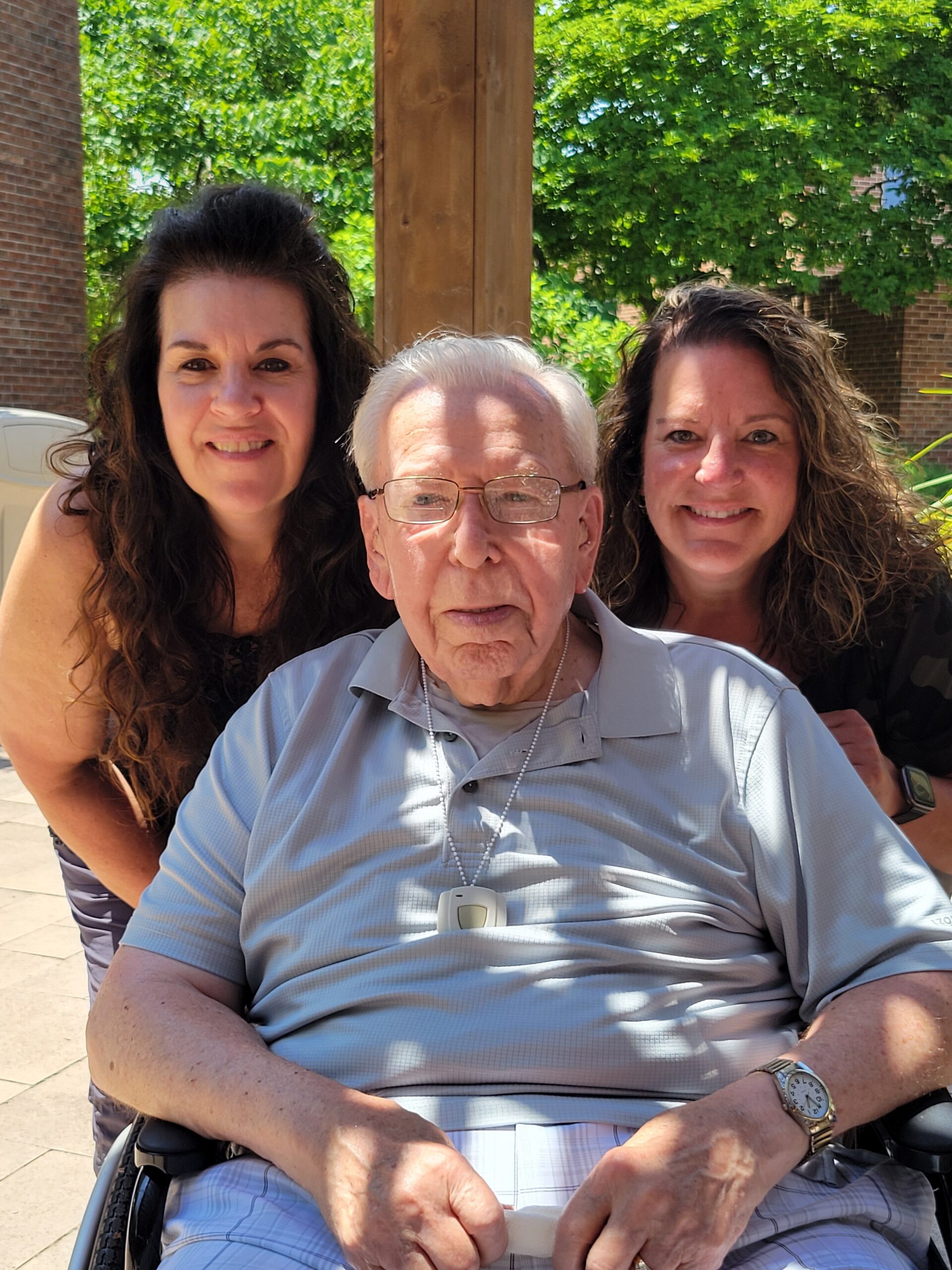 Smiling elderly man in a light blue polo shirt seated in a wheelchair, flanked by two smiling women outdoors under a wooden structure with green trees in the background.