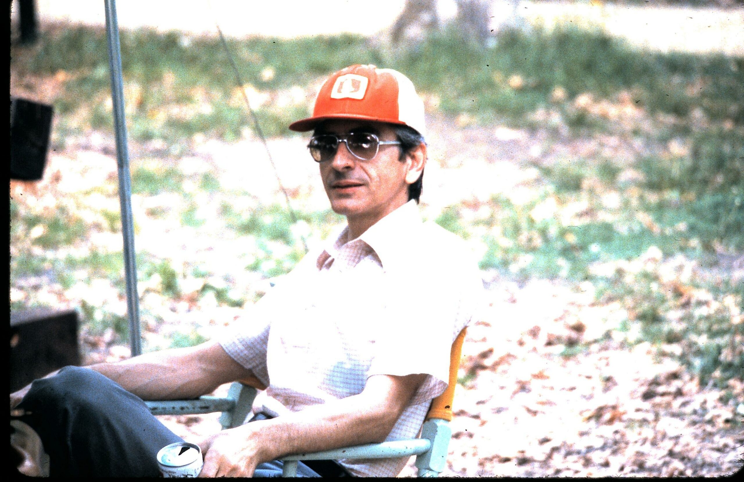 Man sitting in an outdoor chair wearing a red and white baseball cap and sunglasses, with a leafy background.