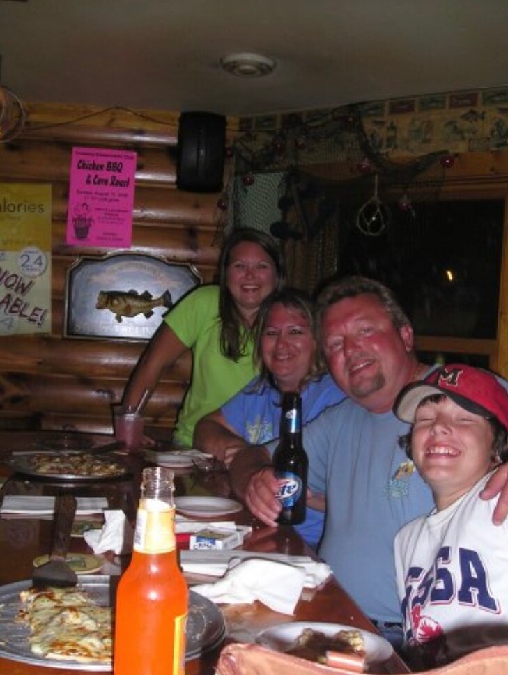 Four friends sit around a wooden restaurant table, smiling at the camera with pizza, drinks, and a bottle of beer visible.