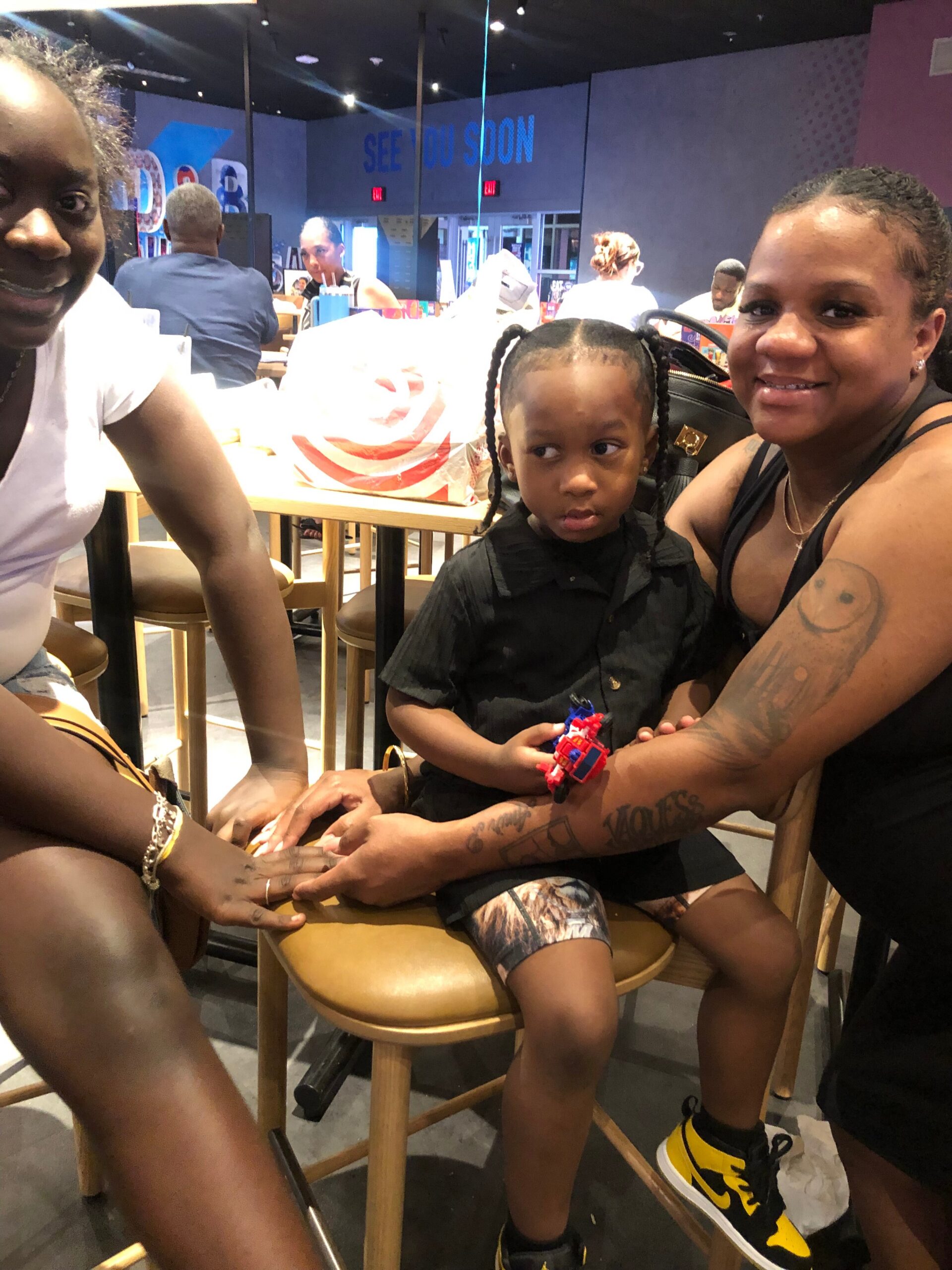 A smiling group of three: a woman in white on the left, a young girl with braided hair in the center wearing a black shirt, and a woman with a black tank top and visible tattoos on the right, sitting on tall chairs in a cafe or store with shelves in the background and a 'SEE YOU SOON' sign behind them.