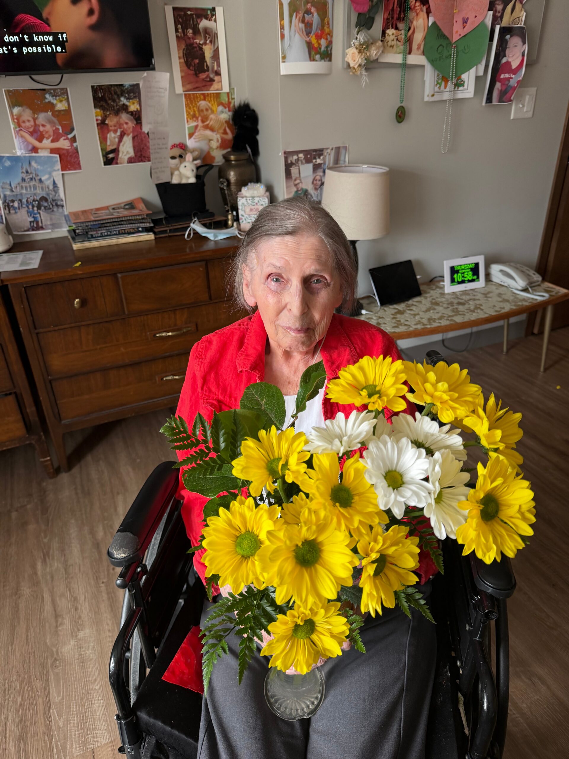 Elderly woman in a wheelchair smiling at the camera, wearing a red top, holding a bouquet of yellow and white flowers in a homey room with photos on the wall behind.