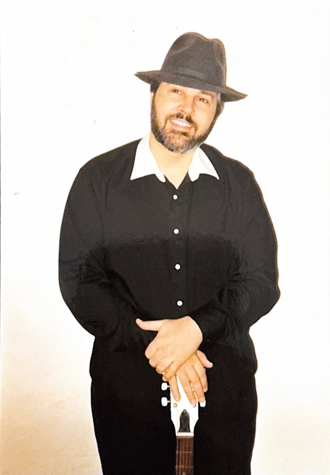 Man in a black button-down shirt and wide-brimmed hat, smiling, holding the neck of an acoustic guitar against a light background