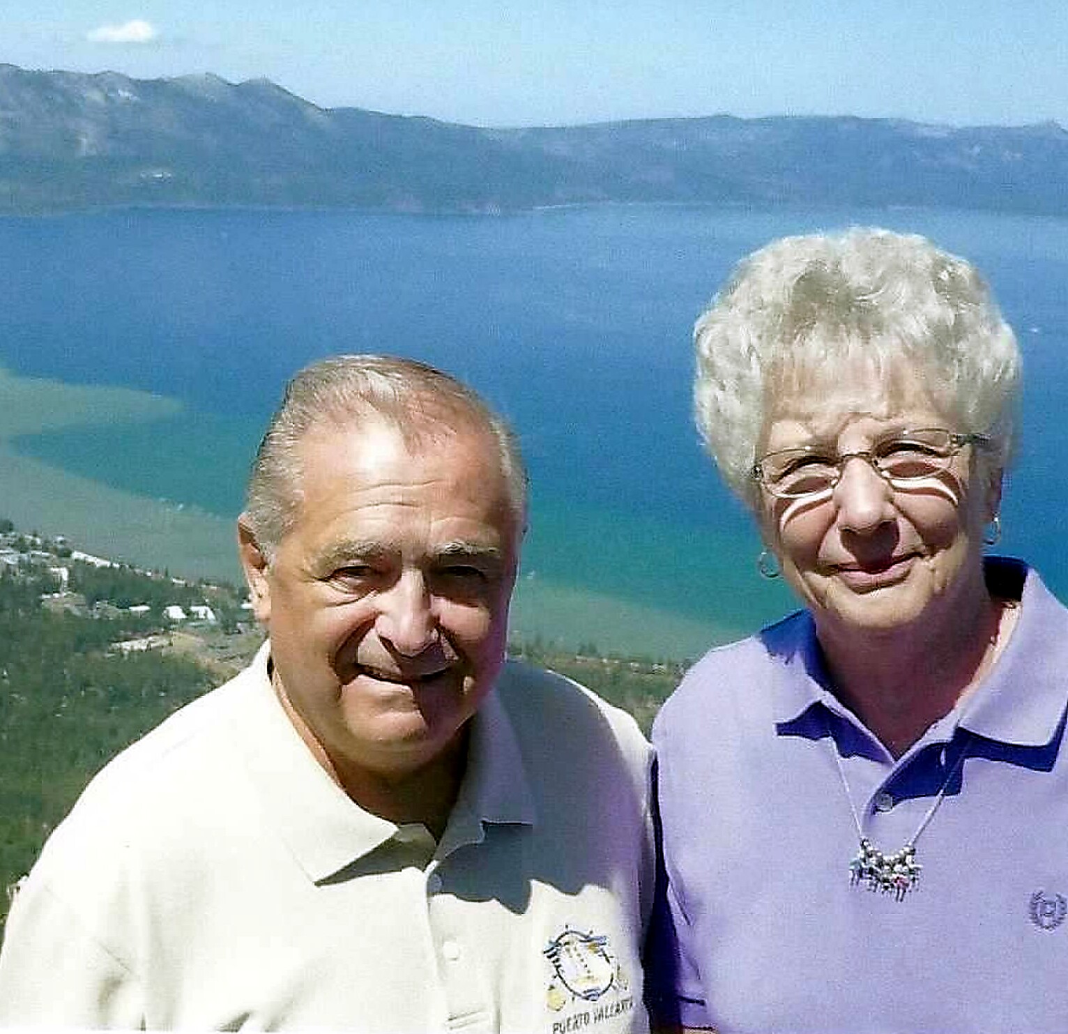 Smiling older couple posing for a photo with a blue lake and distant mountains behind them on a sunny day, near the coast.