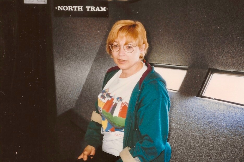 Portrait of a woman with short blond hair and round glasses, seated inside a tram wearing a teal jacket and a colorful graphic T-shirt with a belt visible.