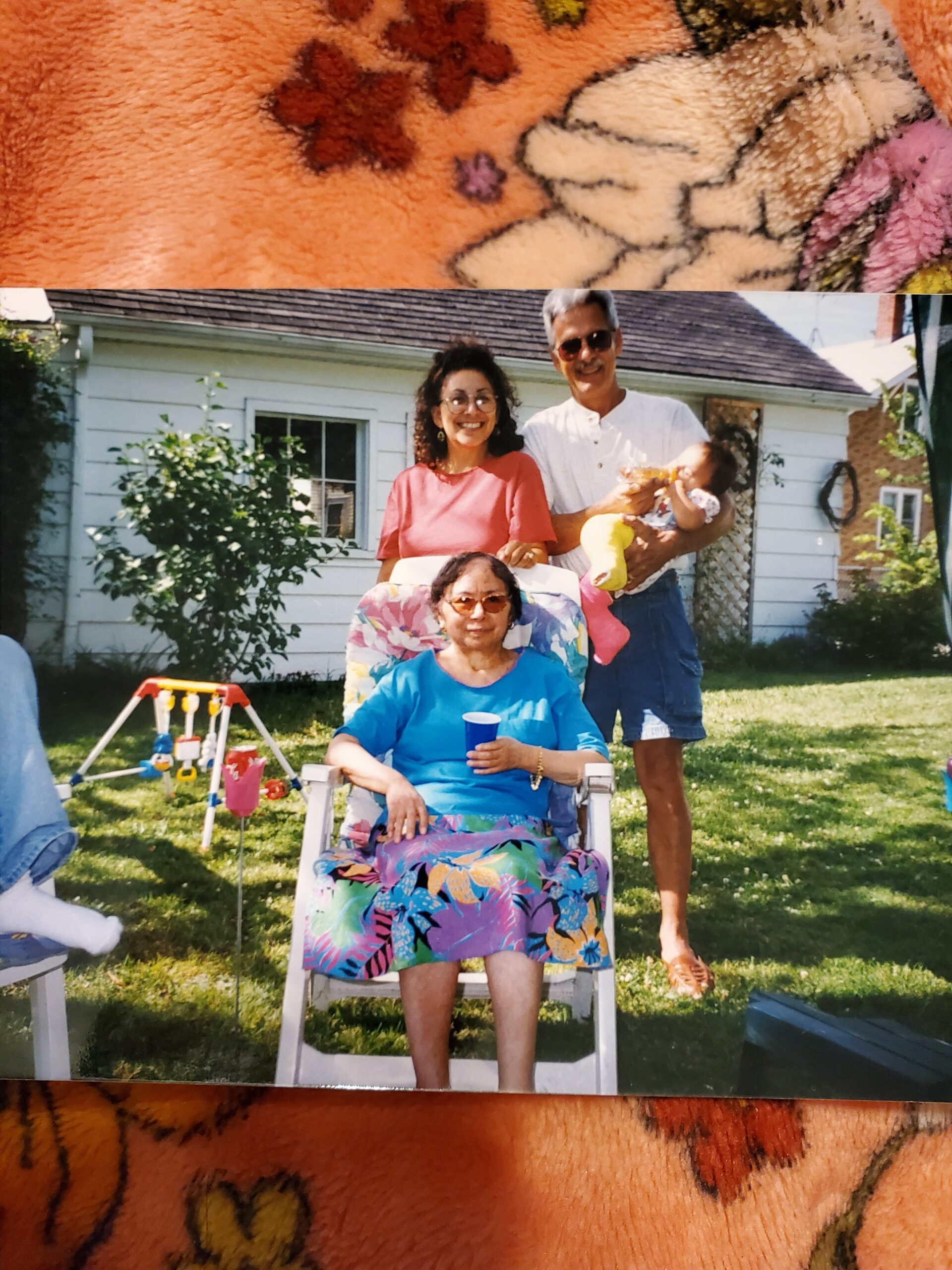 Smiling family in a backyard: an elderly woman in a blue floral dress sits in a white chair while a couple stands behind with a baby in their arms.