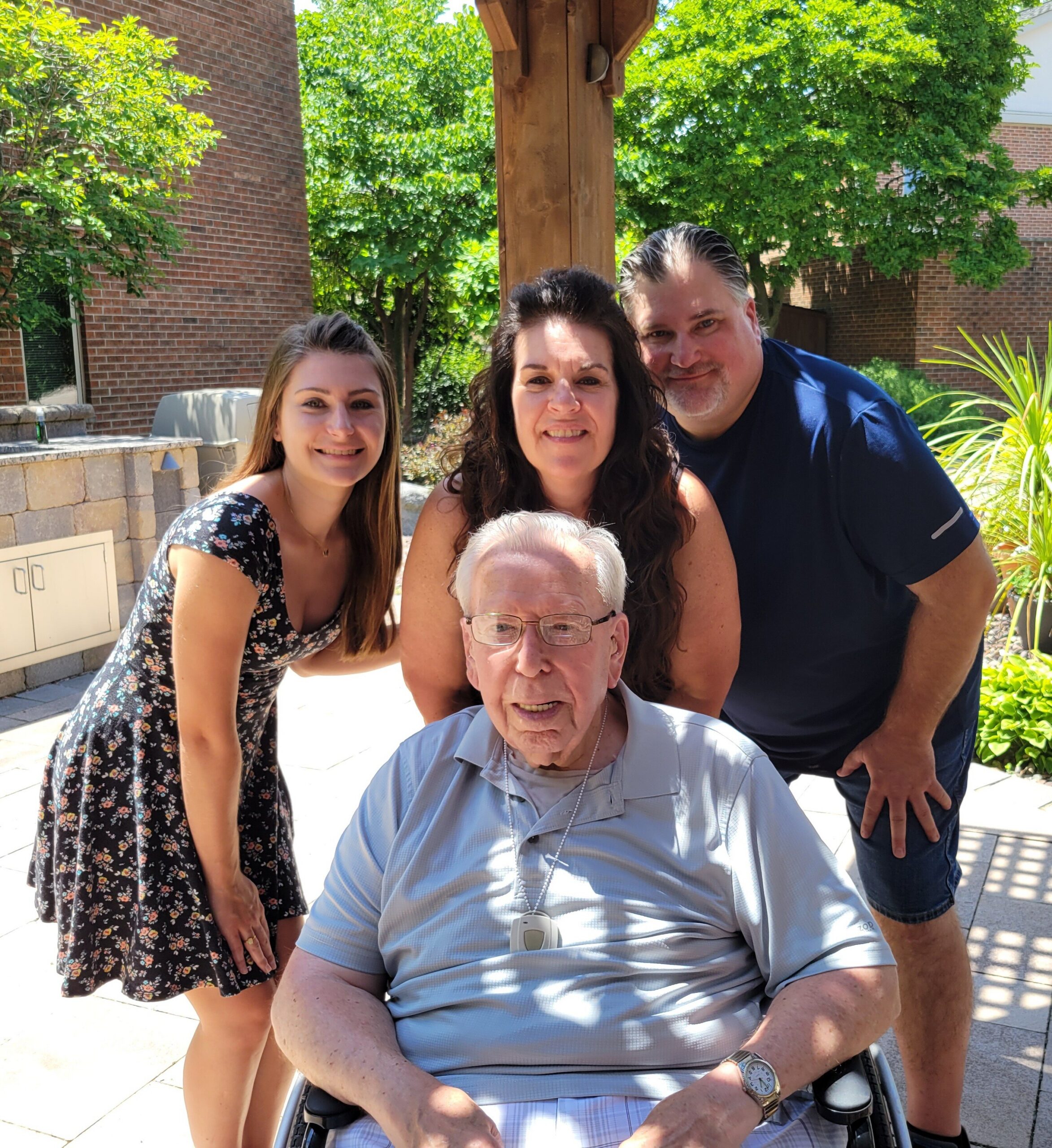 Smiling group of four adults posing outdoors on a sunny patio; elderly man in a wheelchair in front, flanked by two women and a man behind him.