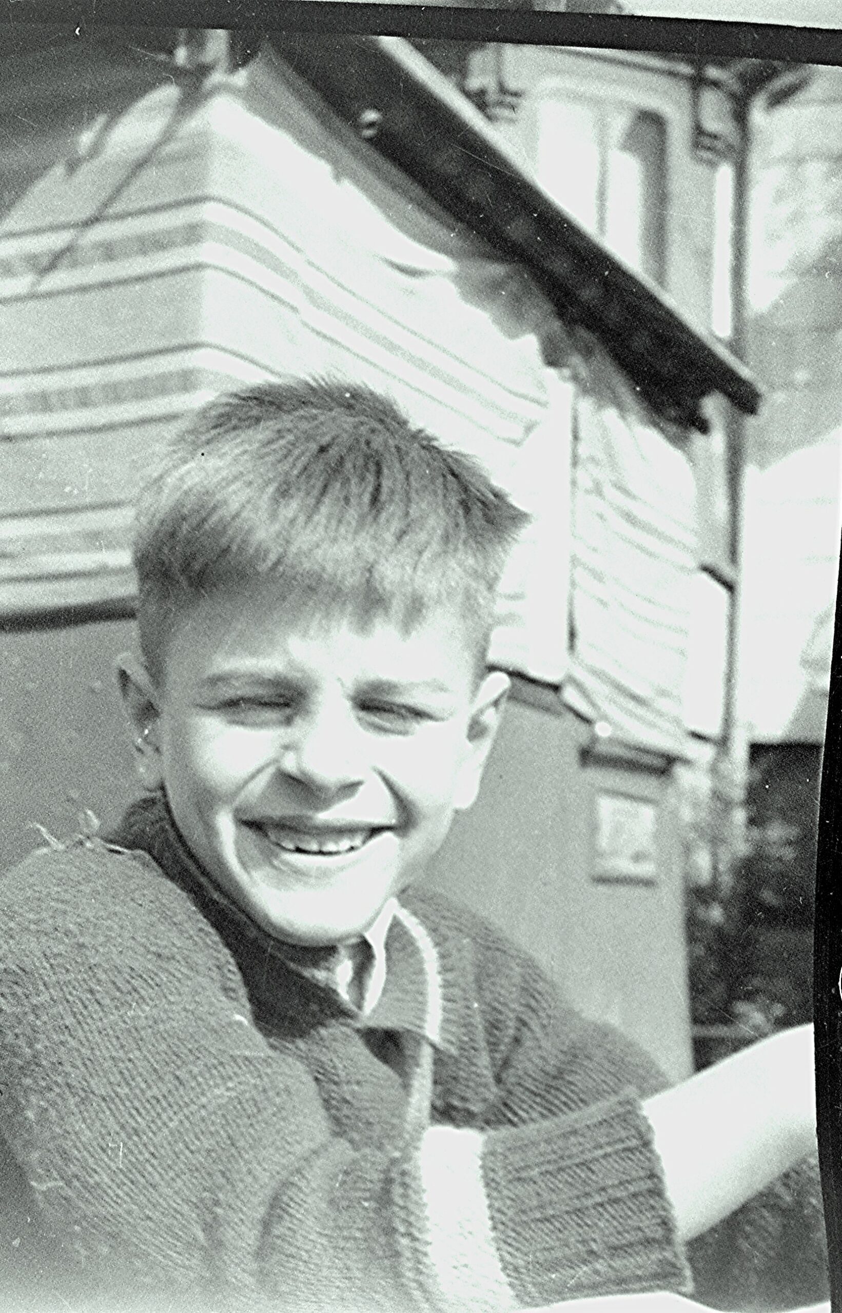 Smiling child with short light hair wearing a sweater, outdoors near a striped structure in black-and-white photo