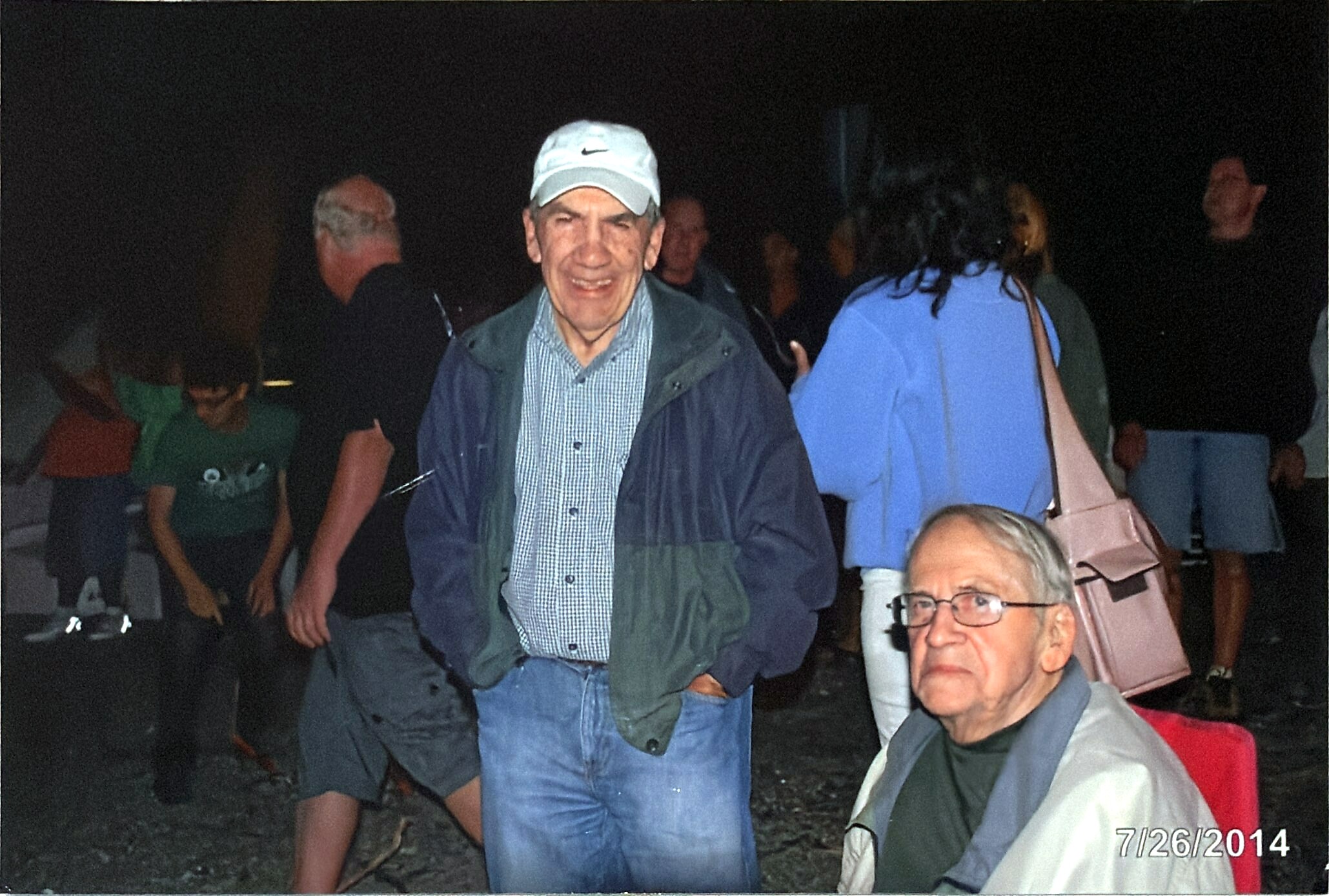 Smiling man wearing a light cap and green jacket stands outdoors at night among a group of people; date stamp visible in bottom right reads 7/26/2014.