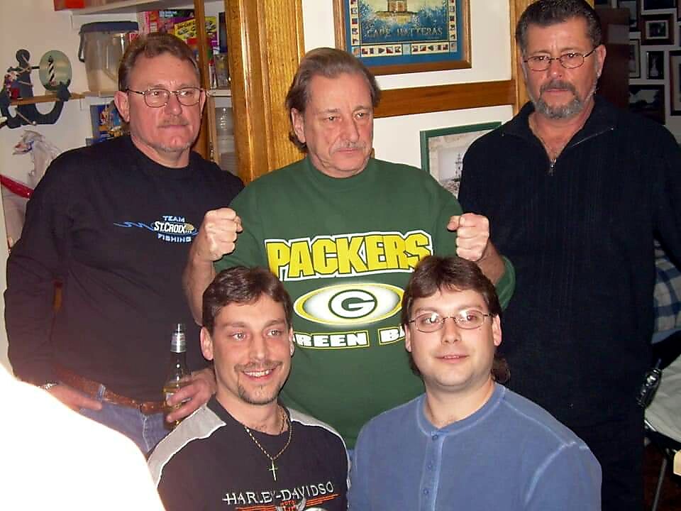 Five men pose for a casual group photo indoors; the center man wears a green Packers shirt. Nearby, others stand or sit in relaxed poses.