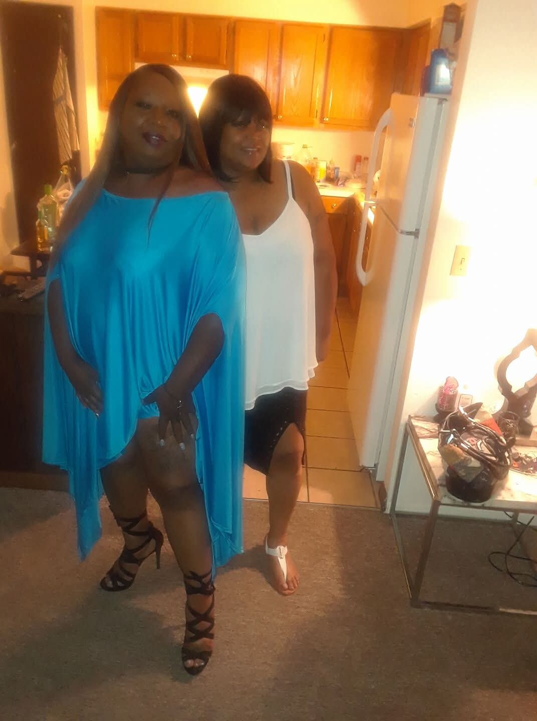 Two women pose in a kitchen; one wears a bright blue dress with strappy heels, the other a white top with black shorts.