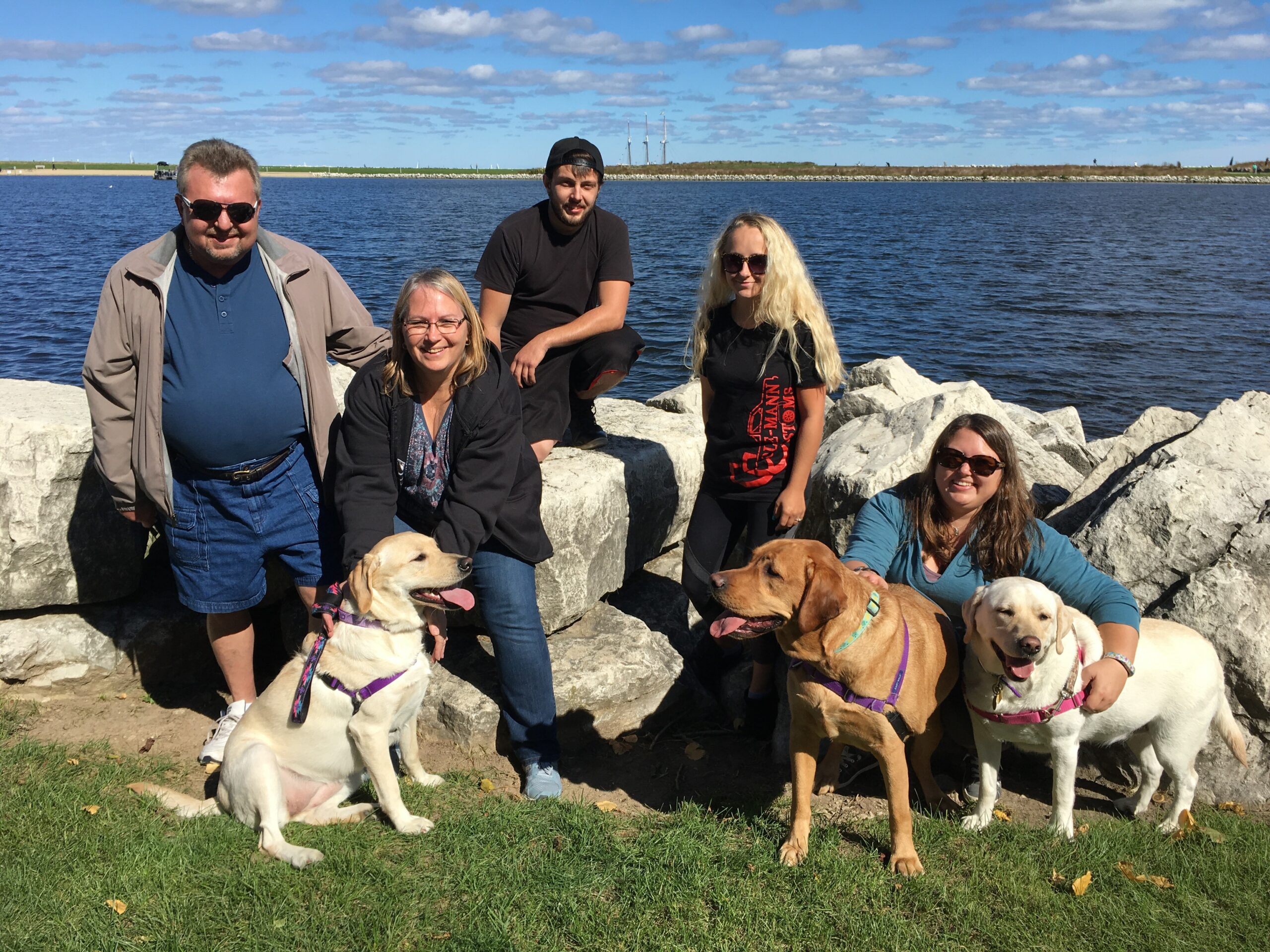 Group of five people and three dogs posing on a rocky shoreline by a large blue lake on a sunny day.