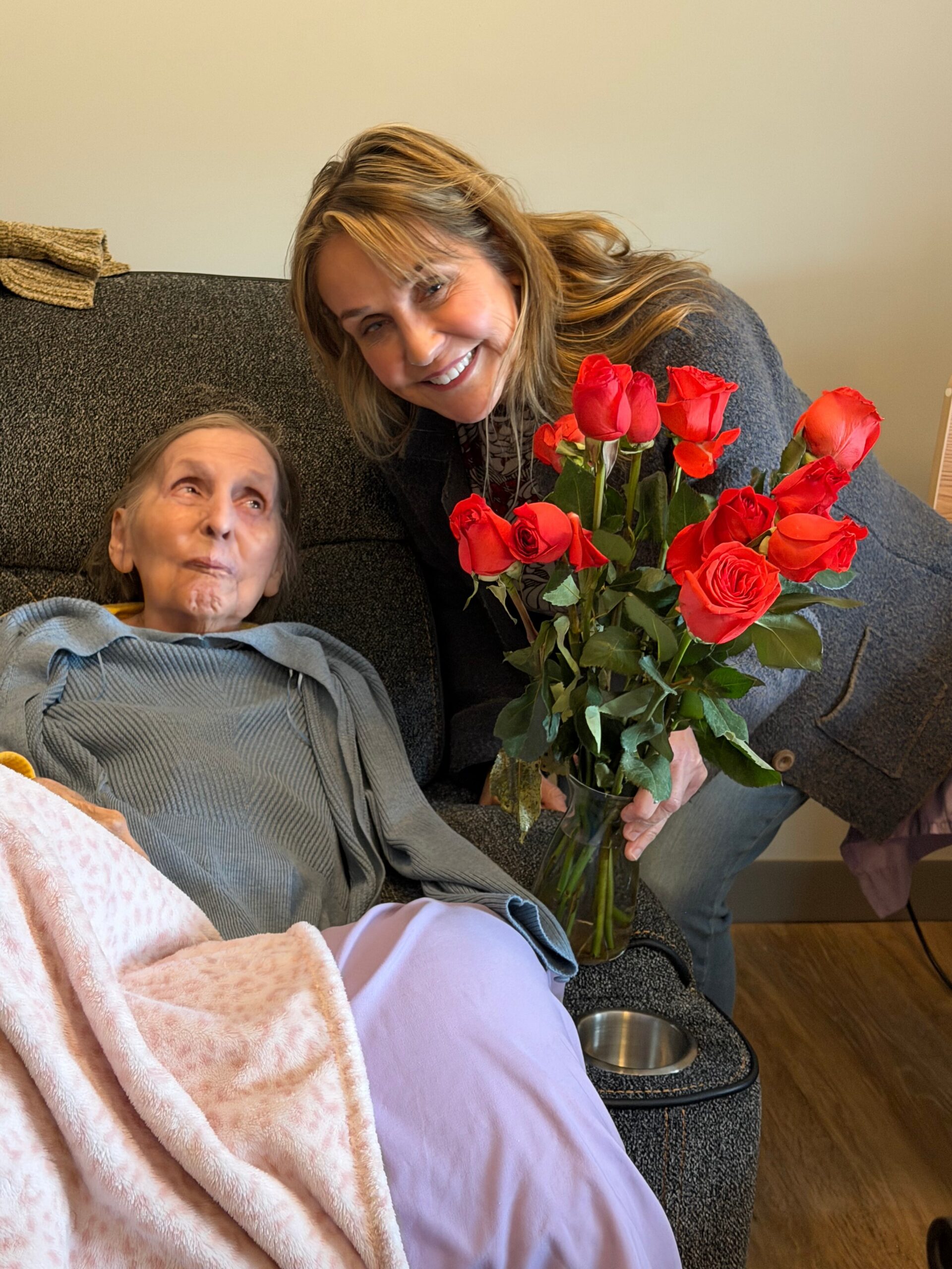 Caregiver presenting a bouquet of red roses to an elderly woman resting on a gray sofa, both smiling warmly.