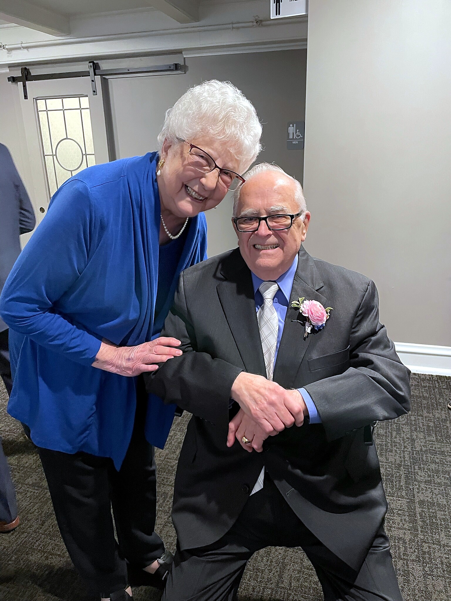 Smiling elderly woman in a blue outfit leaning toward a seated elderly man in a dark suit with a pink corsage, posing together indoors.