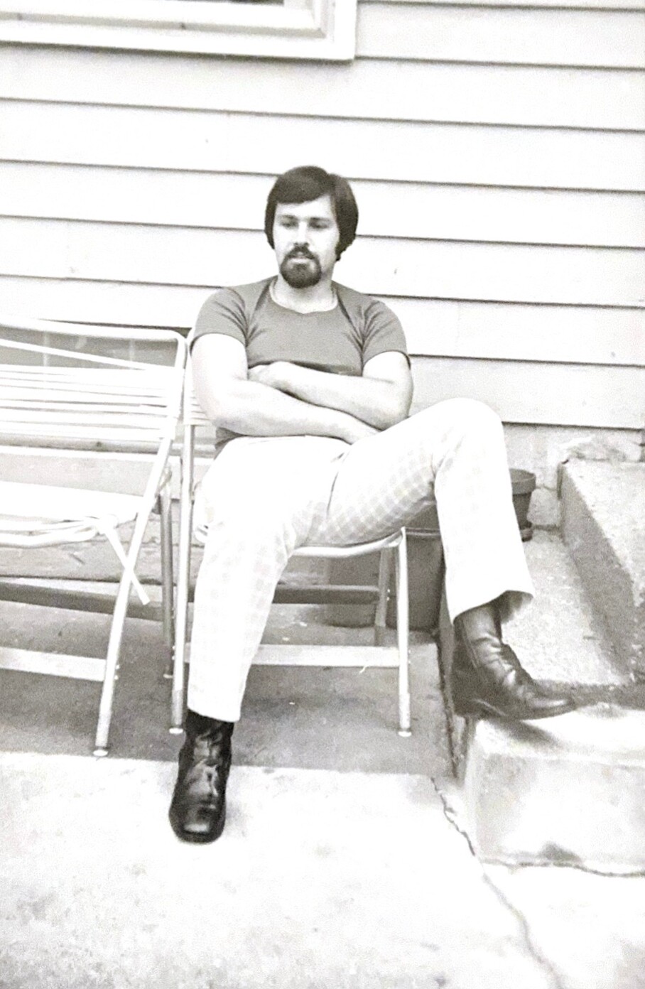 Man with dark hair and mustache sits with arms crossed on a metal outdoor chair beside a siding-walled house.