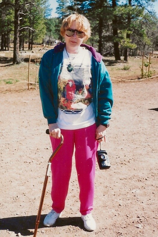 Person standing in a sunny forest clearing, wearing a teal jacket, pink pants, sunglasses, and white sneakers, holding a wooden walking cane in one hand and a small black camera in the other.