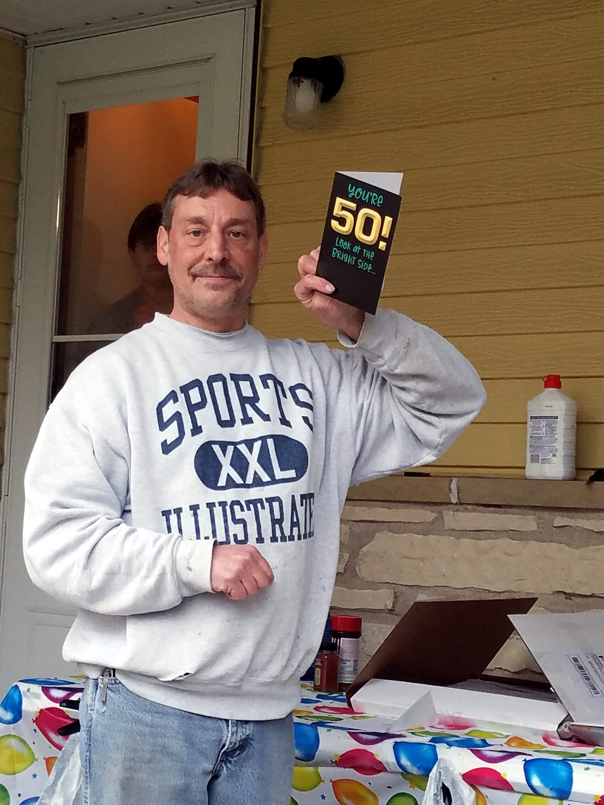Man in a gray sweatshirt smiles while holding a black birthday card that says 'You're 50! Look at the bright side' at an outdoor table party on a wooden porch.