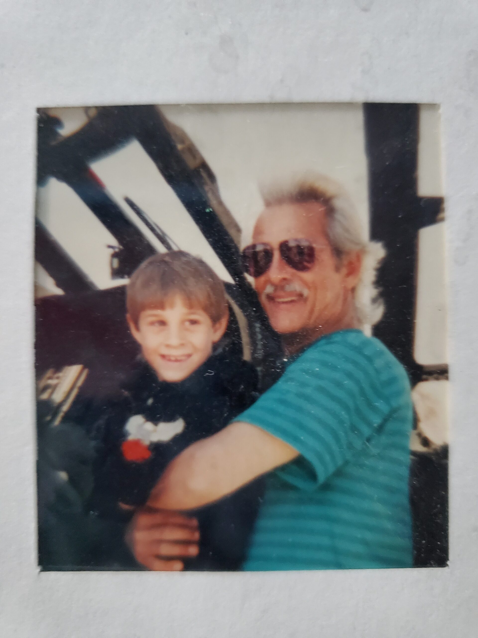 Man with gray hair and sunglasses smiles while holding a young boy in a teal shirt next to him.