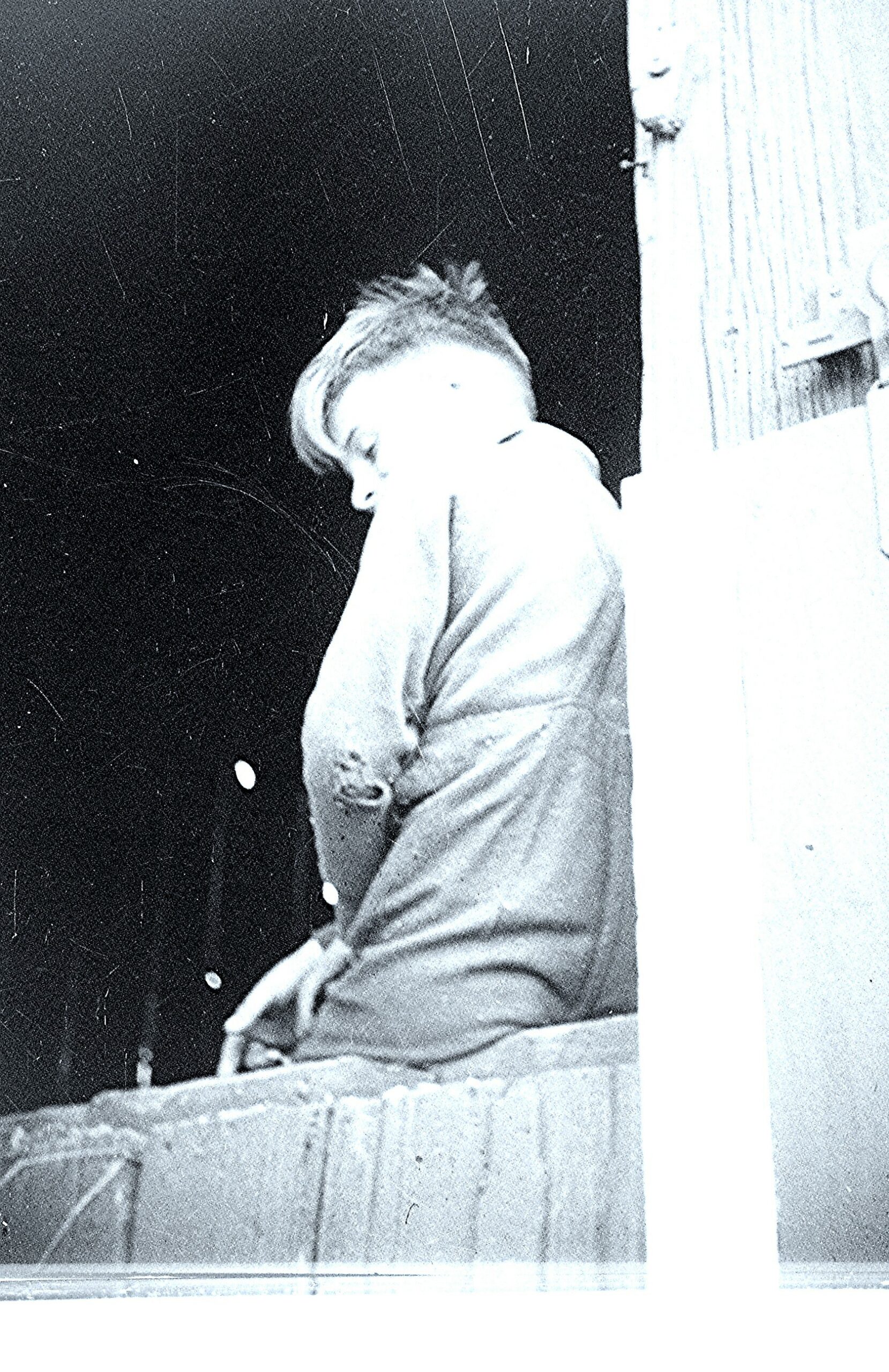 Person seated in profile, leaning against a wooden railing outdoors, head resting on posture, black-and-white photo style
