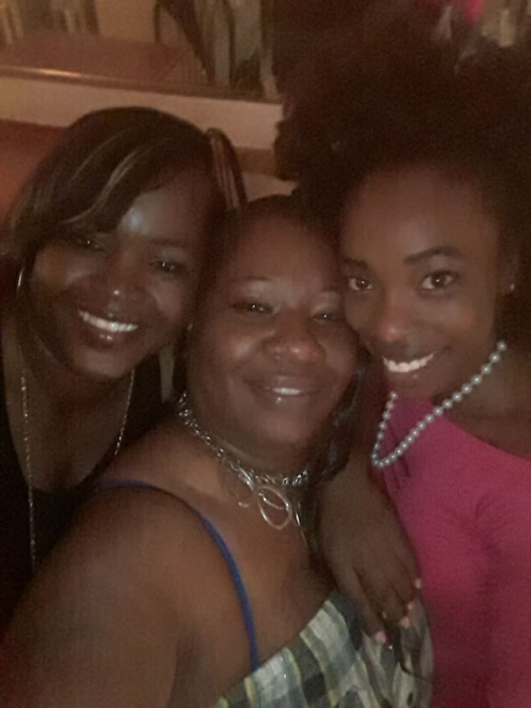 Three women smiling for a close-up selfie, wearing party outfits and jewelry in a dimly lit setting.