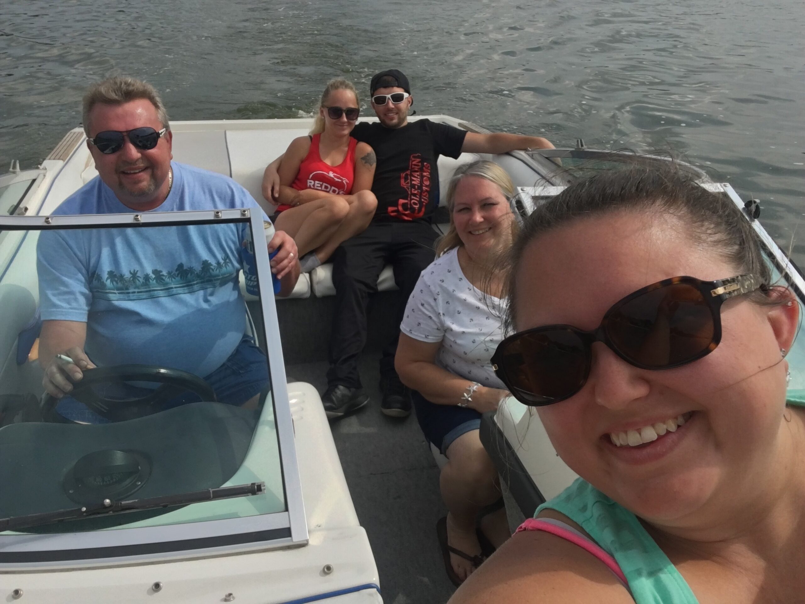 Group of five friends on a boat taking a selfie, smiling and wearing sunglasses, with water in the background.