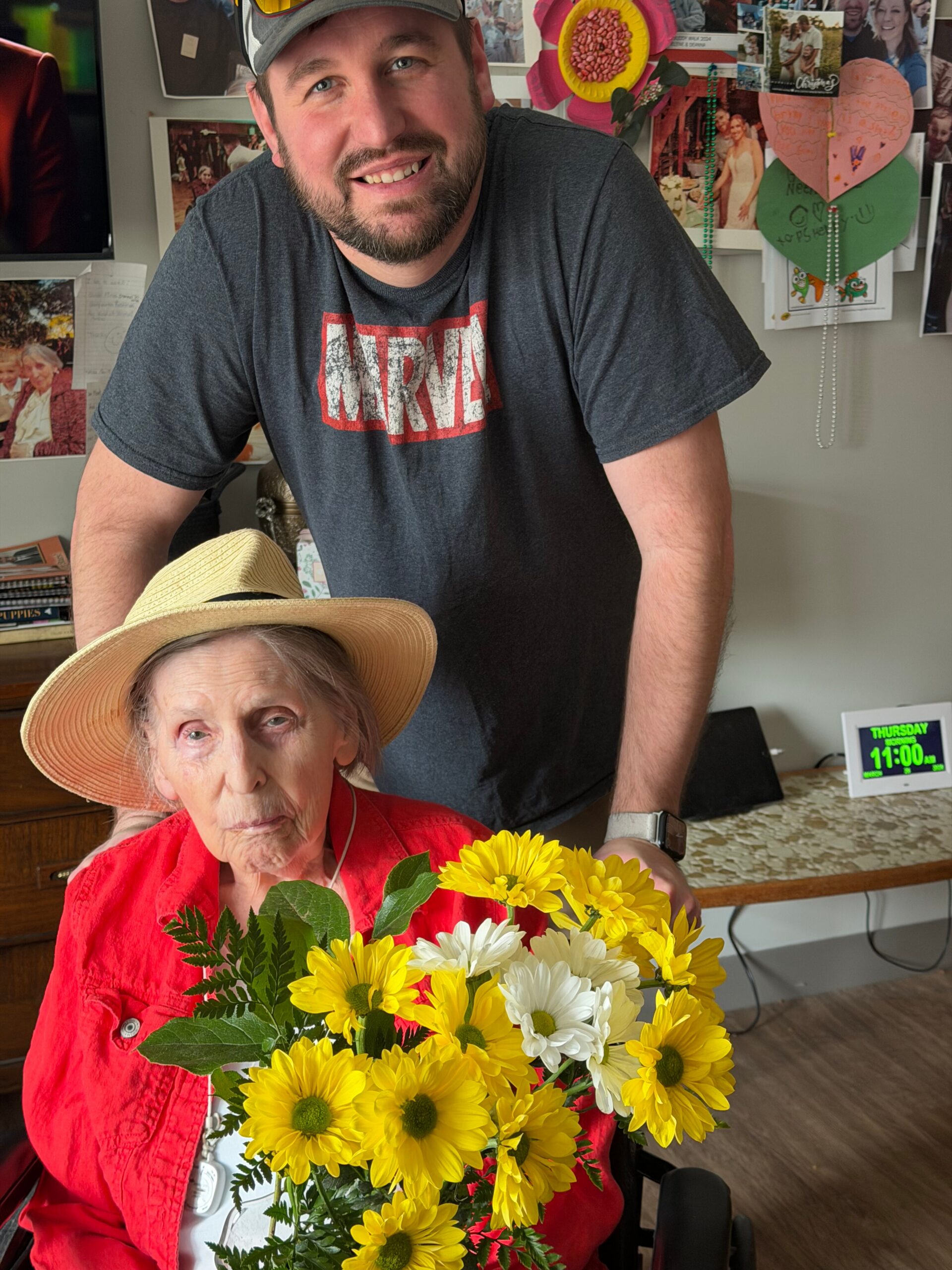Elderly woman in a red blouse in a wheelchair holds a bouquet of yellow and white flowers; a smiling man in a gray Marvel shirt stands behind her.