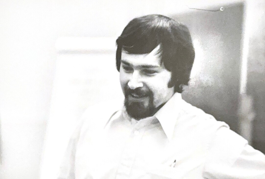 Black-and-white photo of a man with dark hair and a goatee, smiling while looking downward in a light shirt.