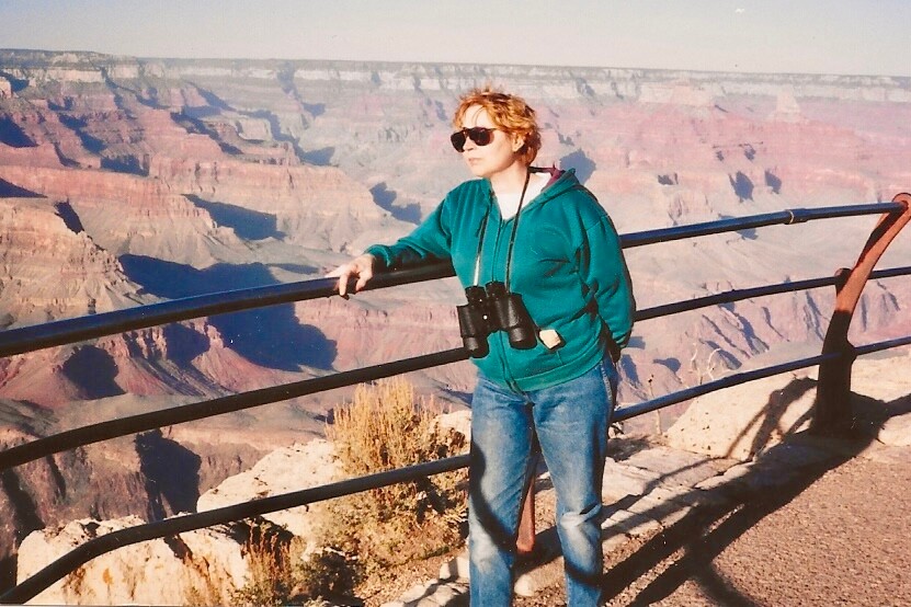 Person in a teal hoodie and sunglasses stands at a railing, binoculars hanging from their neck, overlooking the Grand Canyon.
