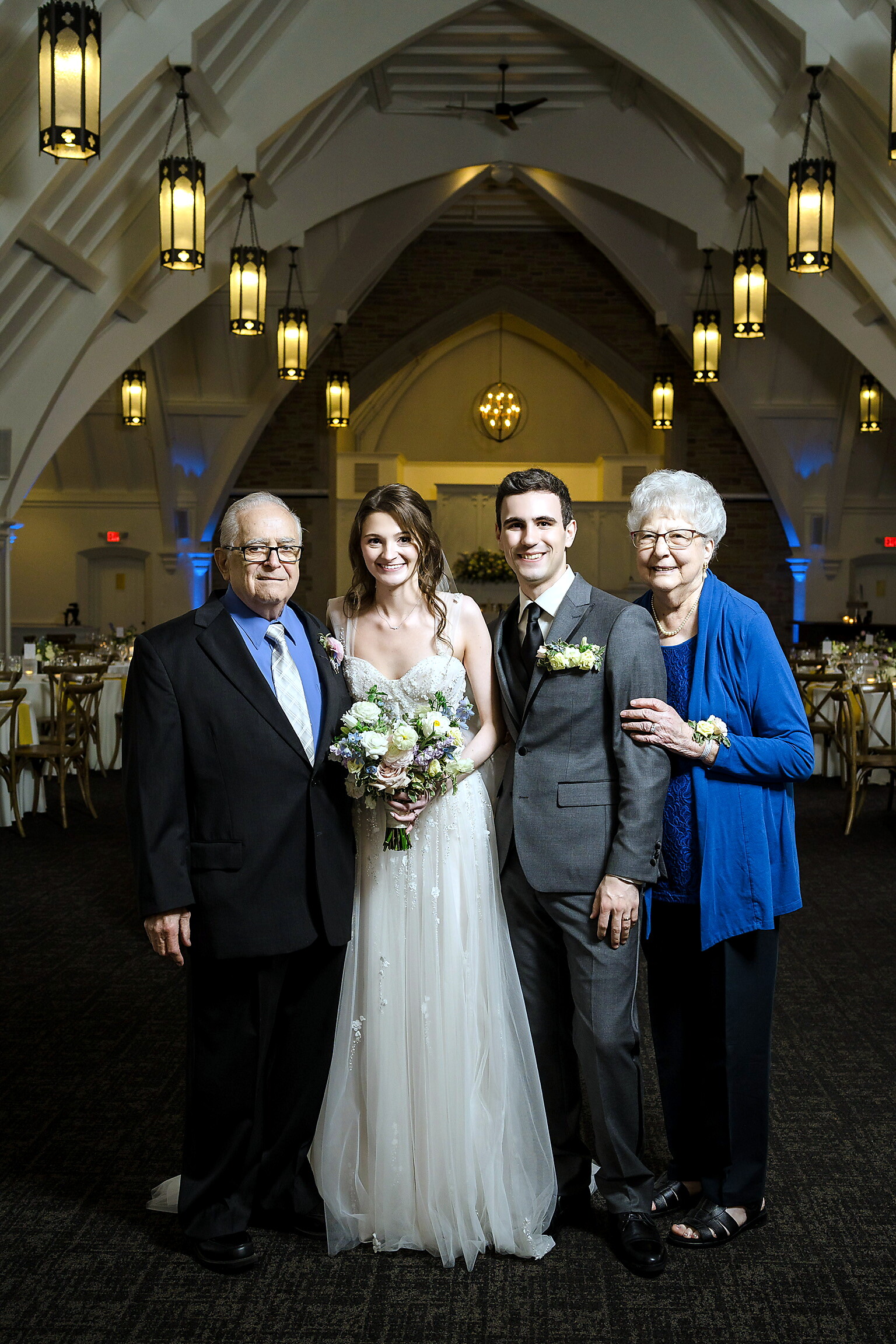 Bride and groom pose with their parents in a decorated wedding venue, smiling for the photo.