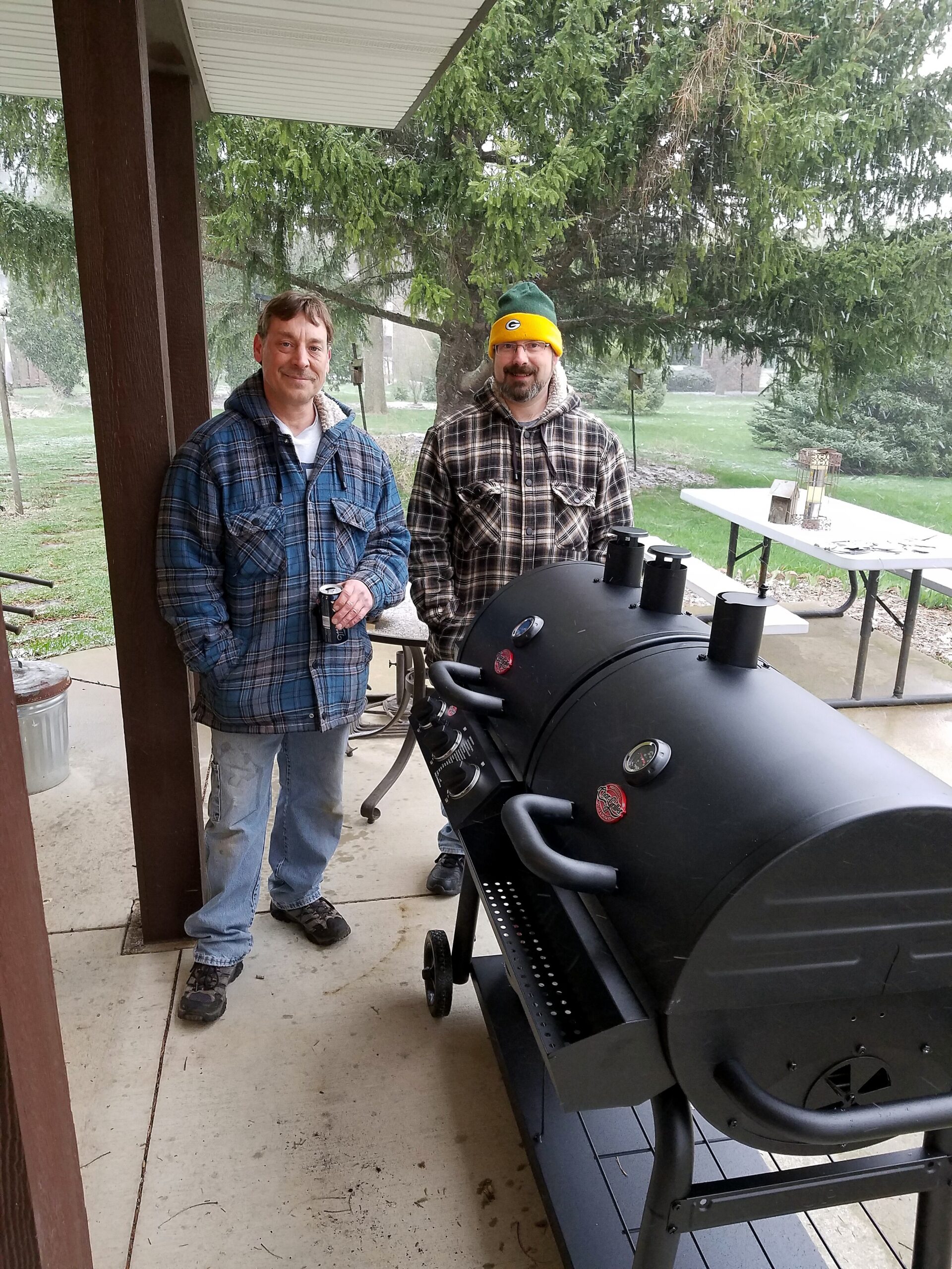 Two men in plaid jackets stand beside a large black outdoor barbecue smoker on a covered patio during light rain; one wears a Green Bay Packers hat.