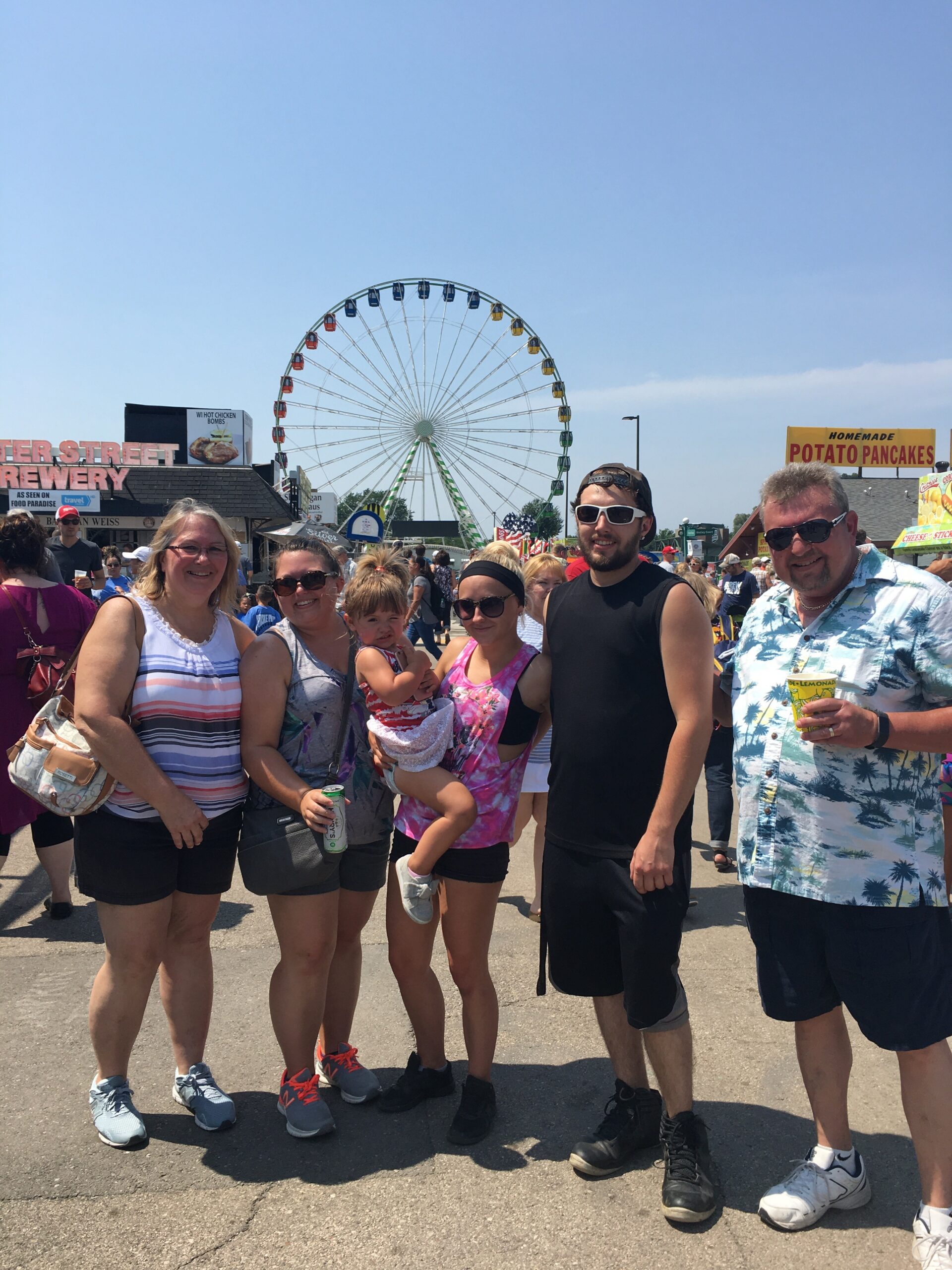 Group of adults and a child posing for a photo at a sunny fairground, with a large Ferris wheel in the background.